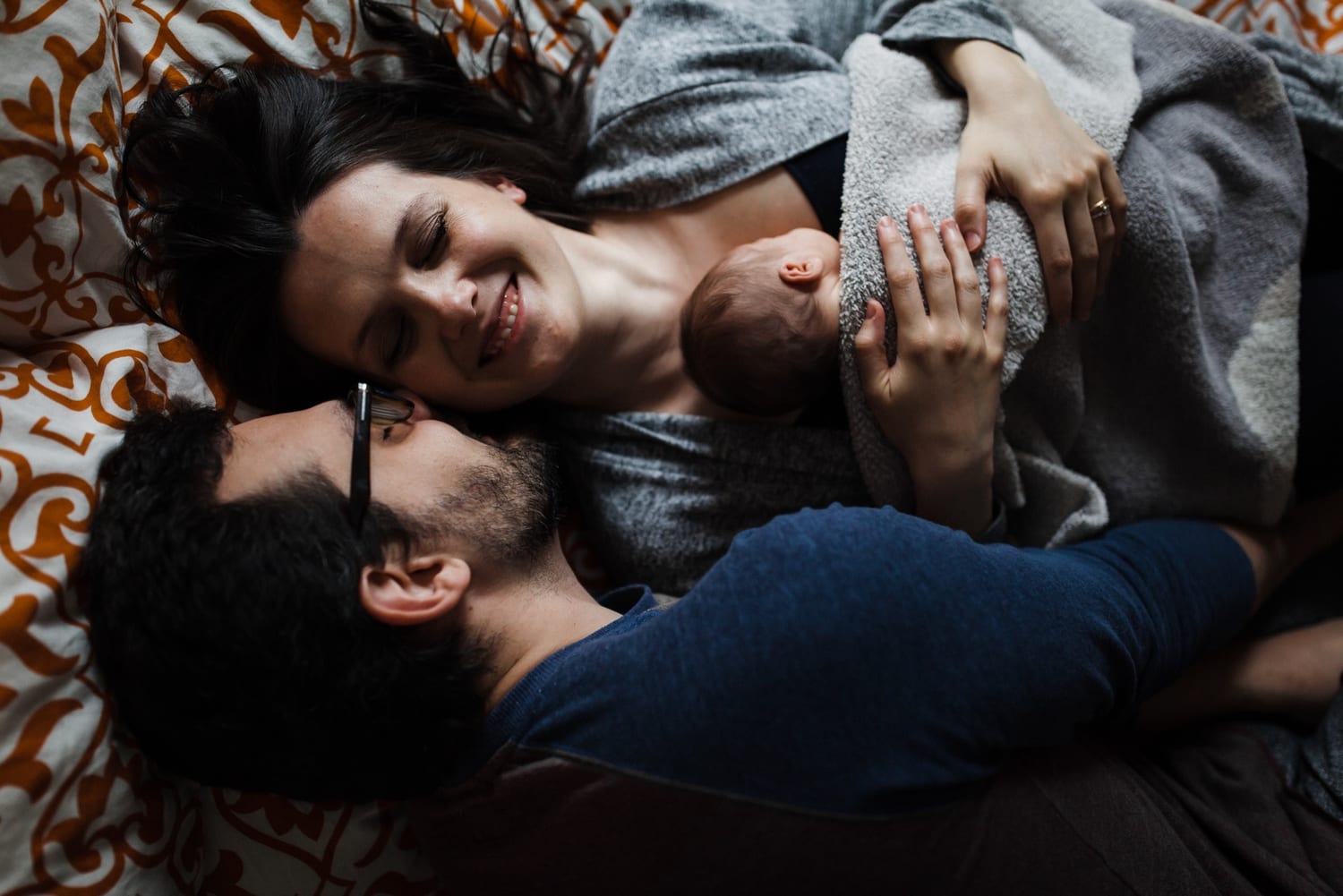 Mom and dad lie on the bed smiling, newborn tucked between them