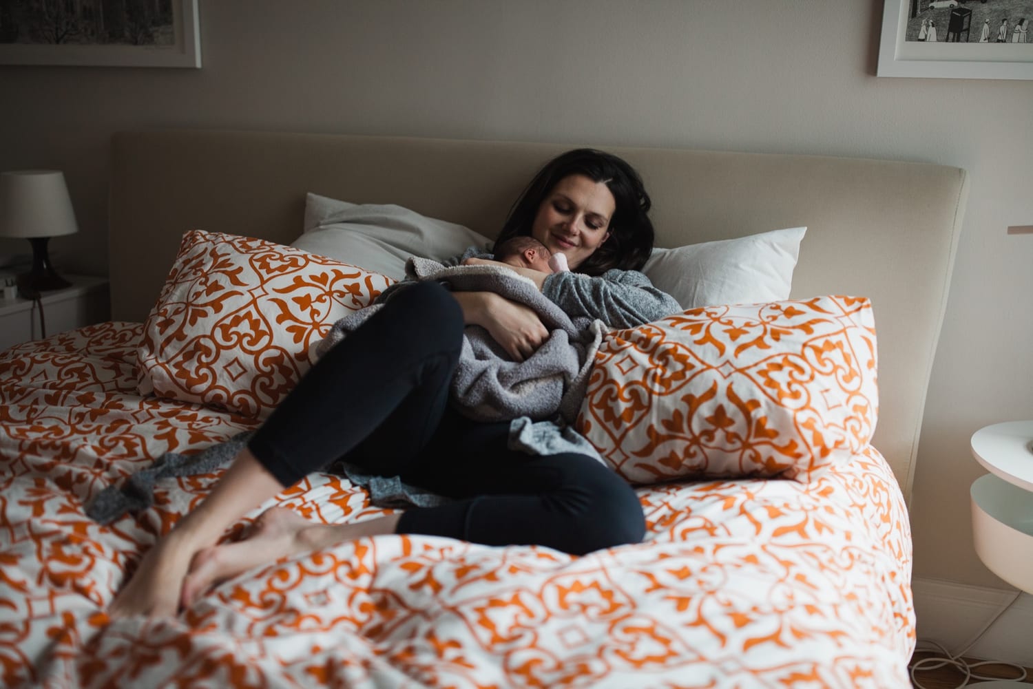 Mom relaxes against the pillows cradling her swaddled newborn on the bed