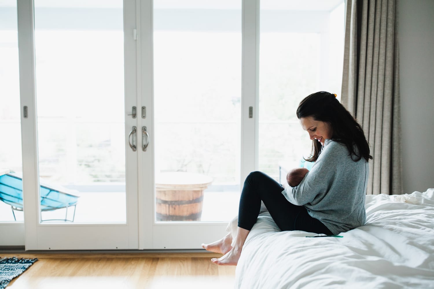 Mom sits on the edge of the bed holding her newborn by French doors