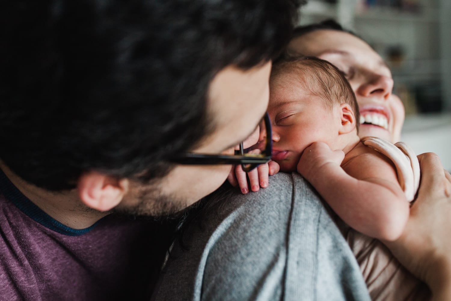 Dad leans in to kiss the newborn asleep on smiling mom's shoulder, in-home newborn photography San Francisco