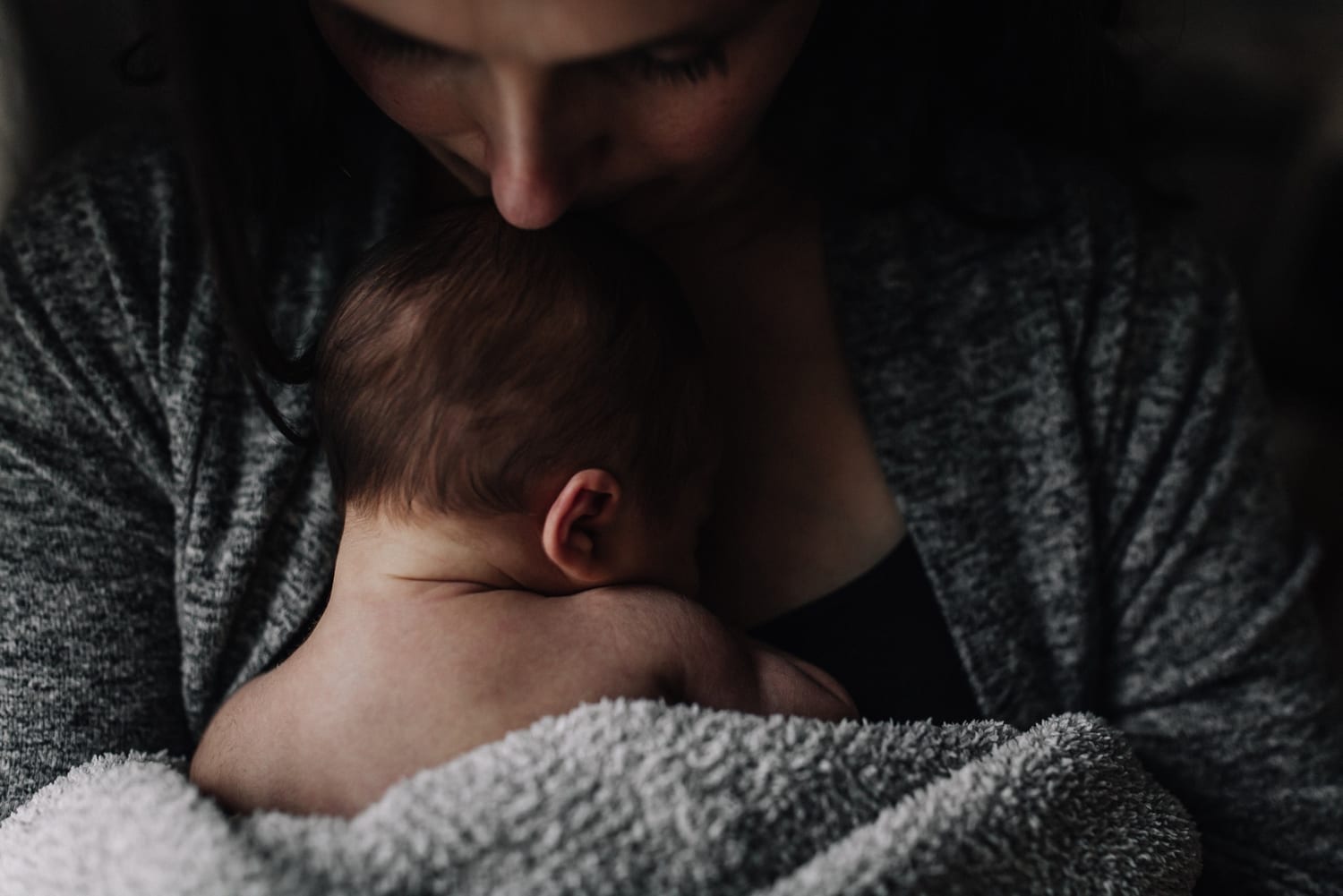 Mom presses a kiss to her newborn's head in soft low light