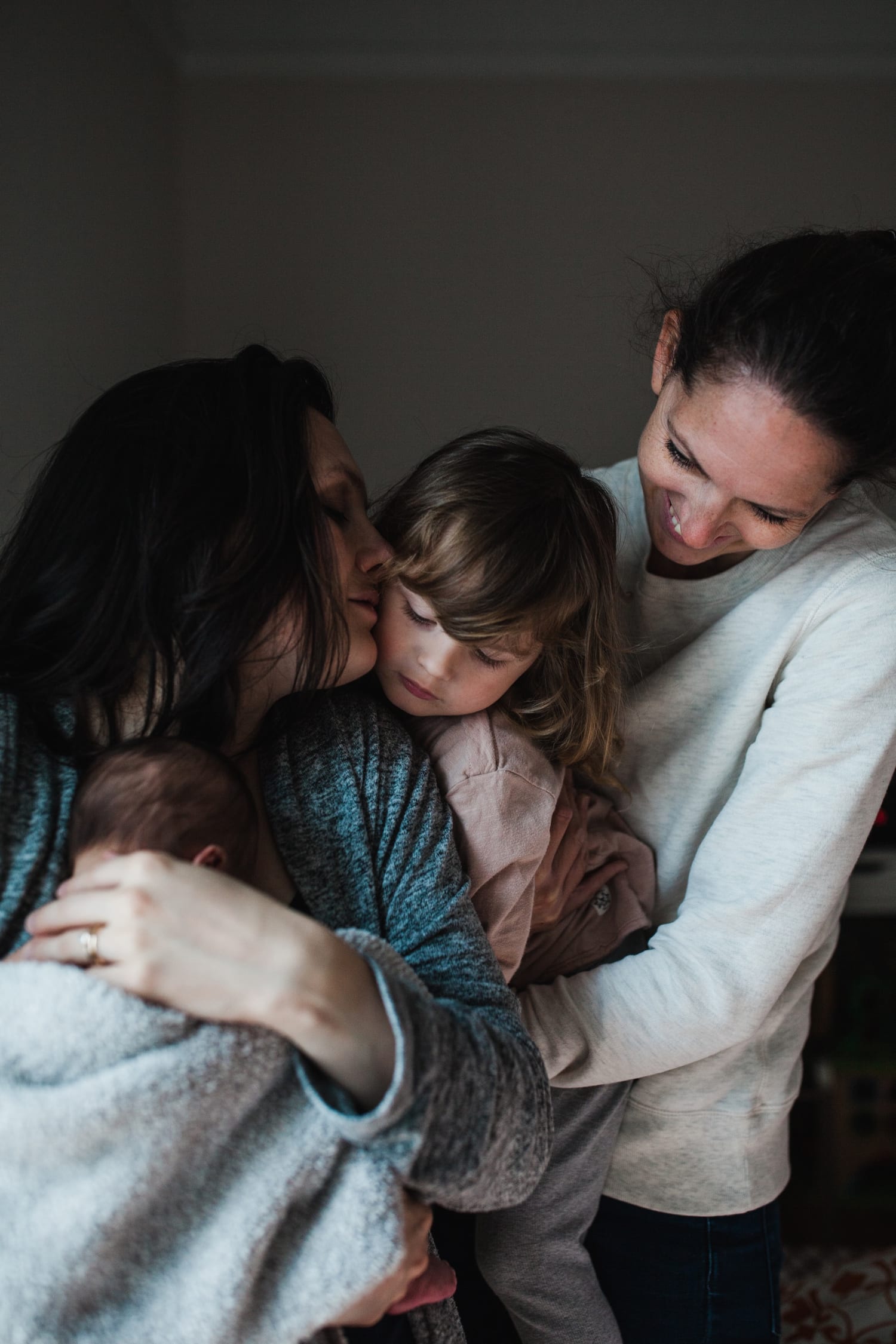 Mom kisses big sister's cheek while holding newborn, wrapped in a family hug