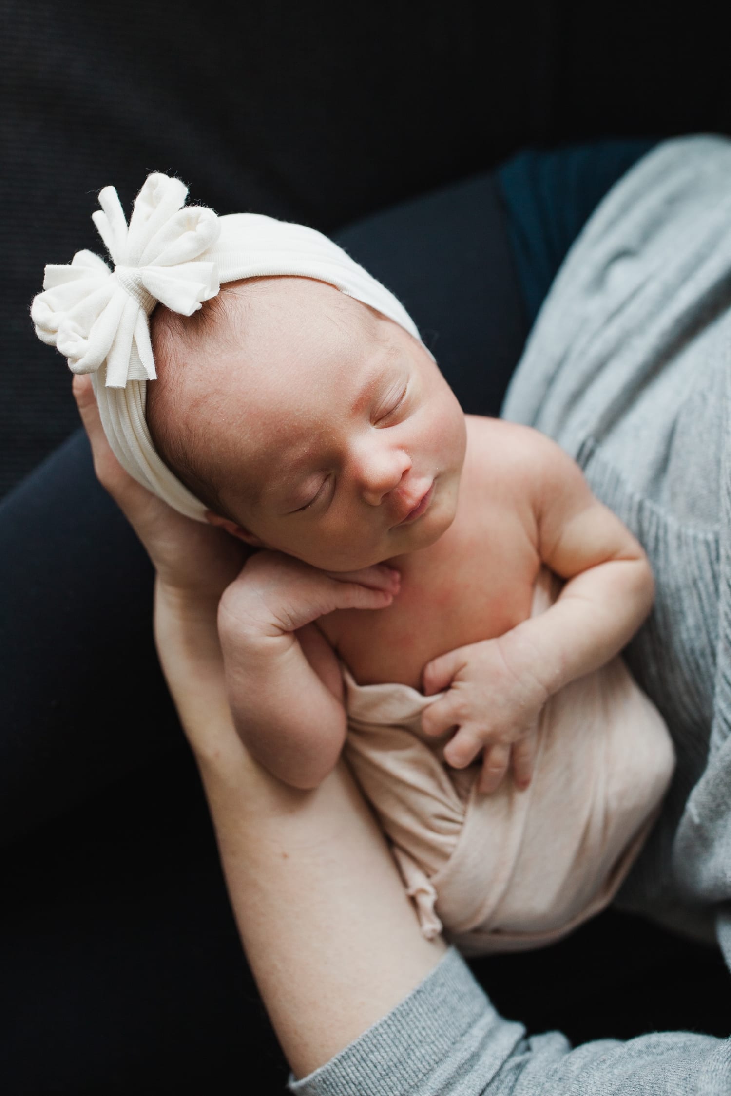 Sleeping newborn wearing a white bow headband, held in mom's hands