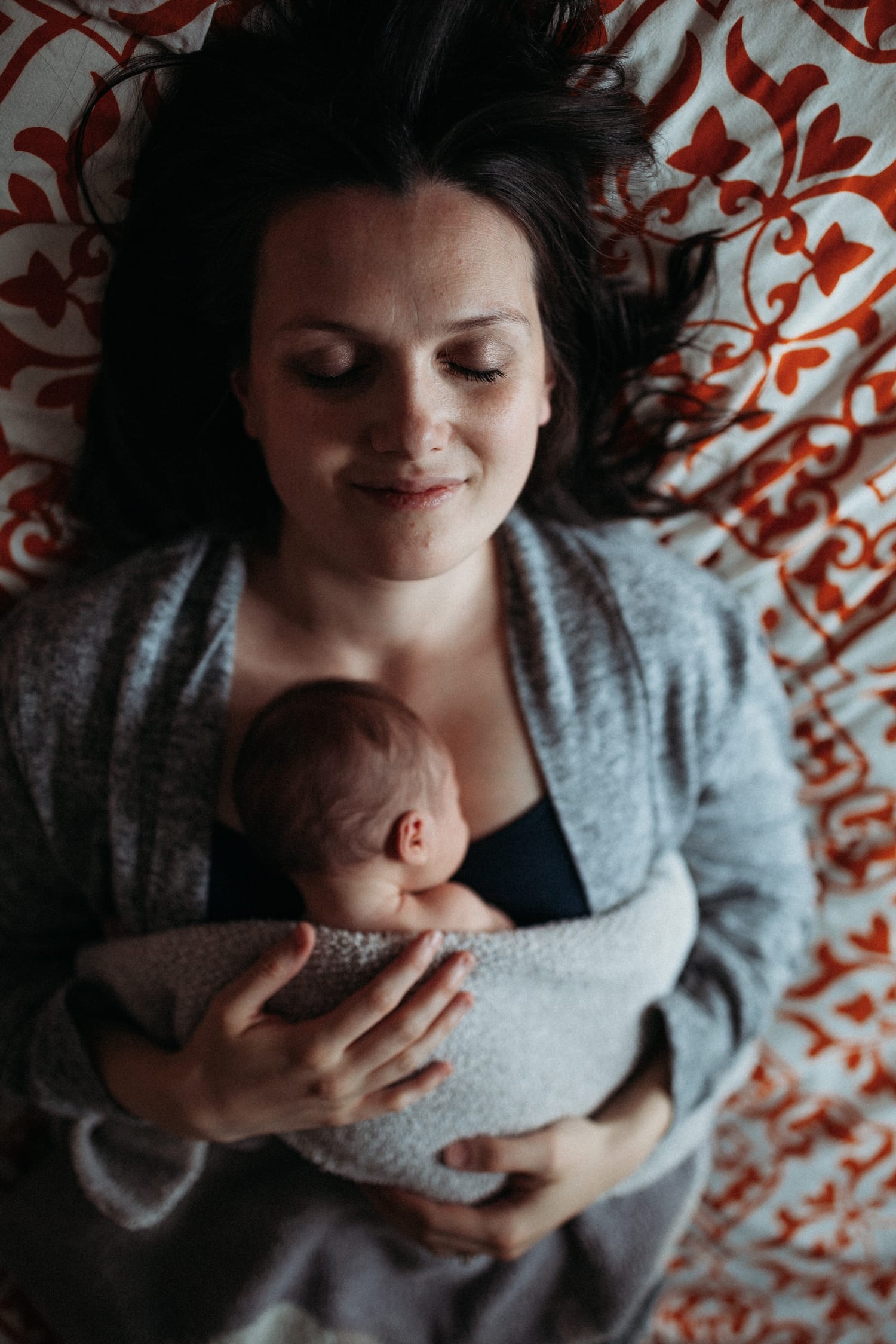 Mom lies on a patterned duvet smiling, sleeping newborn bundled on her chest