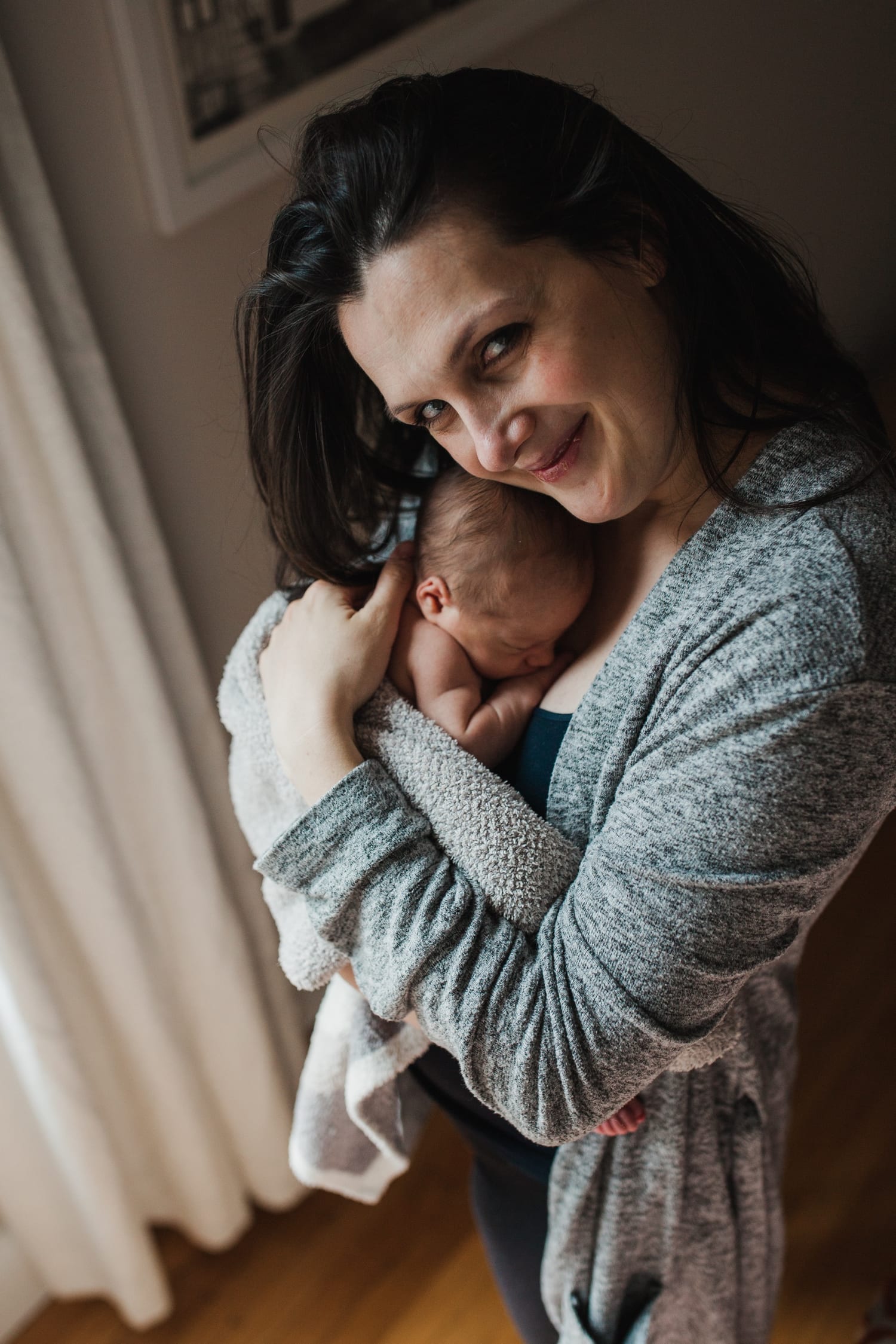 Mom smiles at the camera while hugging her newborn close by the window