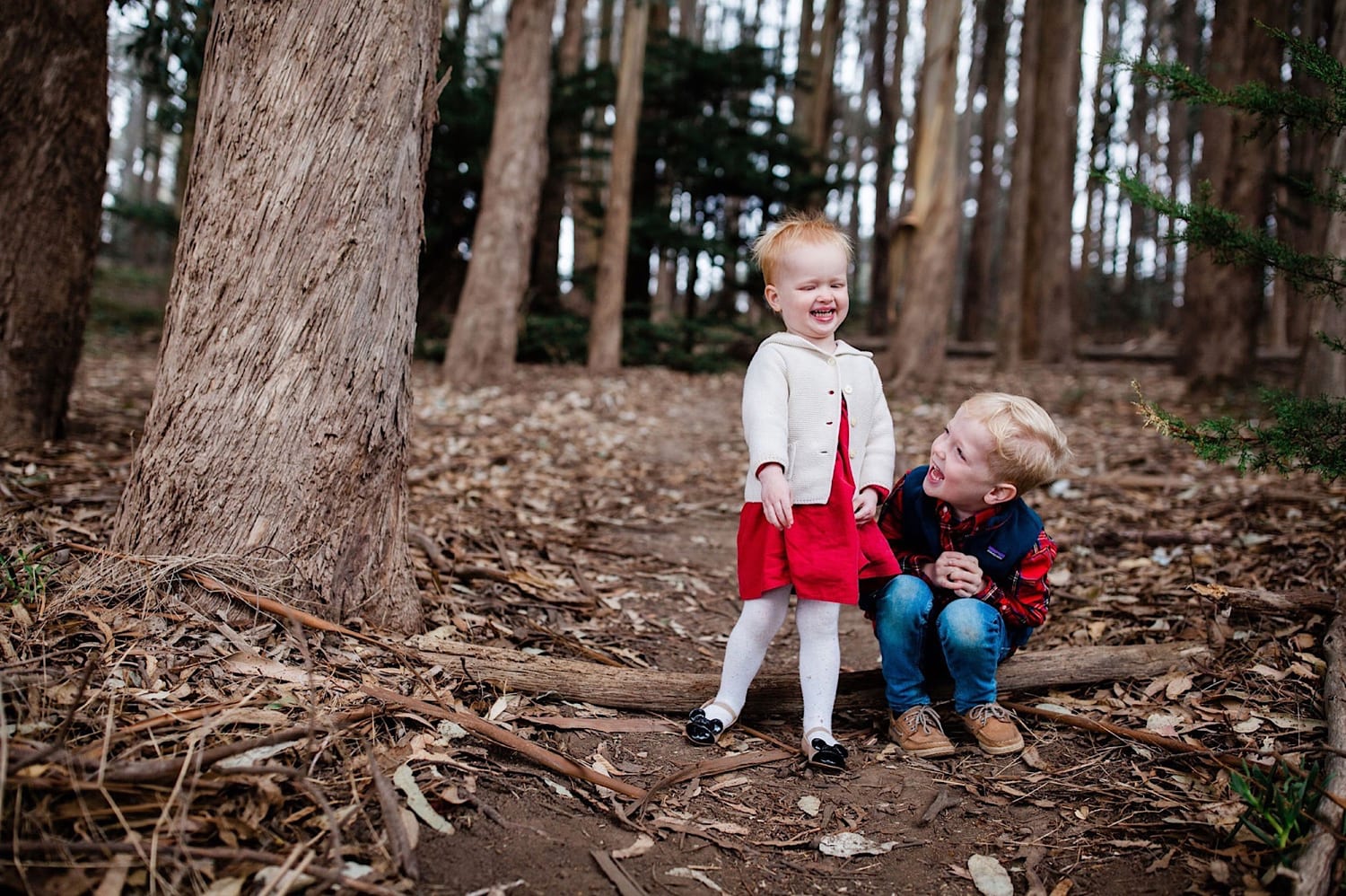 Toddler girl in red dress laughs with her brother beside a tree