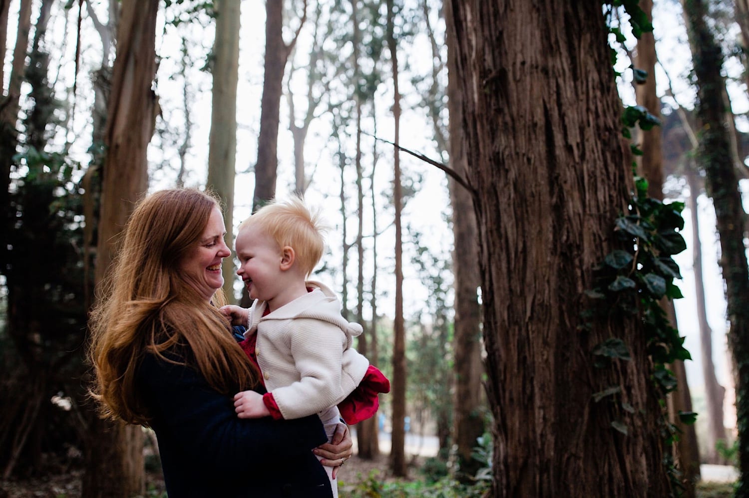 Mom holds her laughing toddler girl face to face in the forest