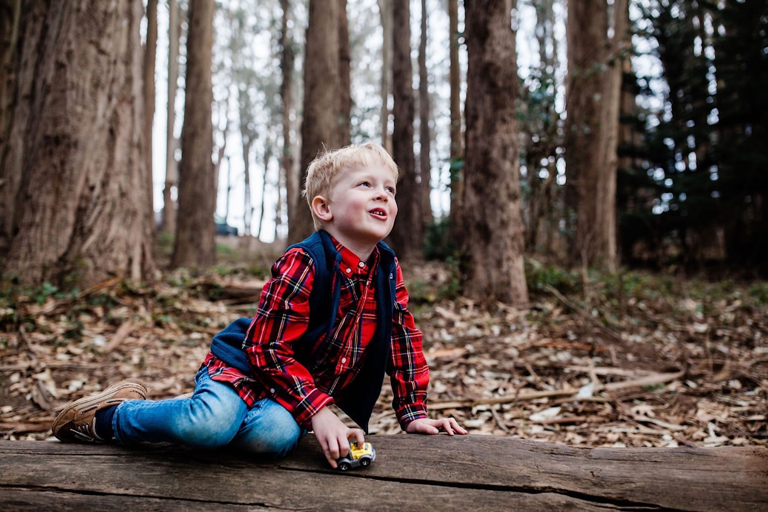 Boy plays with a toy car on a log, looking up and smiling