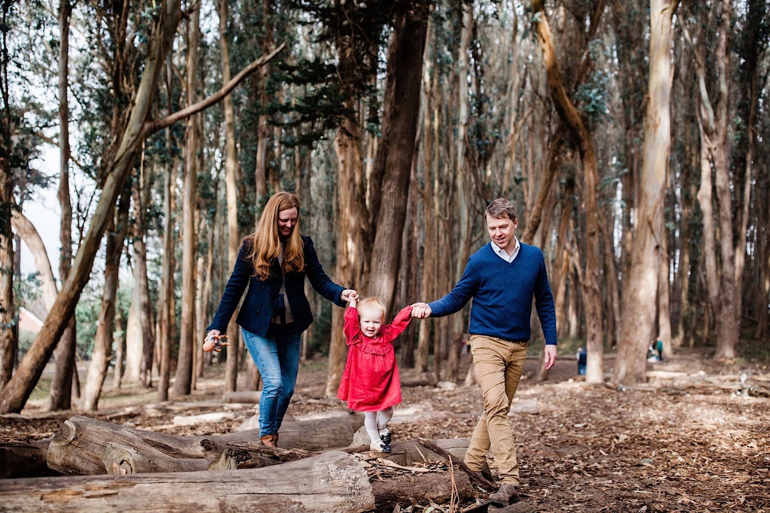 Parents hold toddler's hands as she walks a log in her red dress, Lovers Lane San Francisco family photography