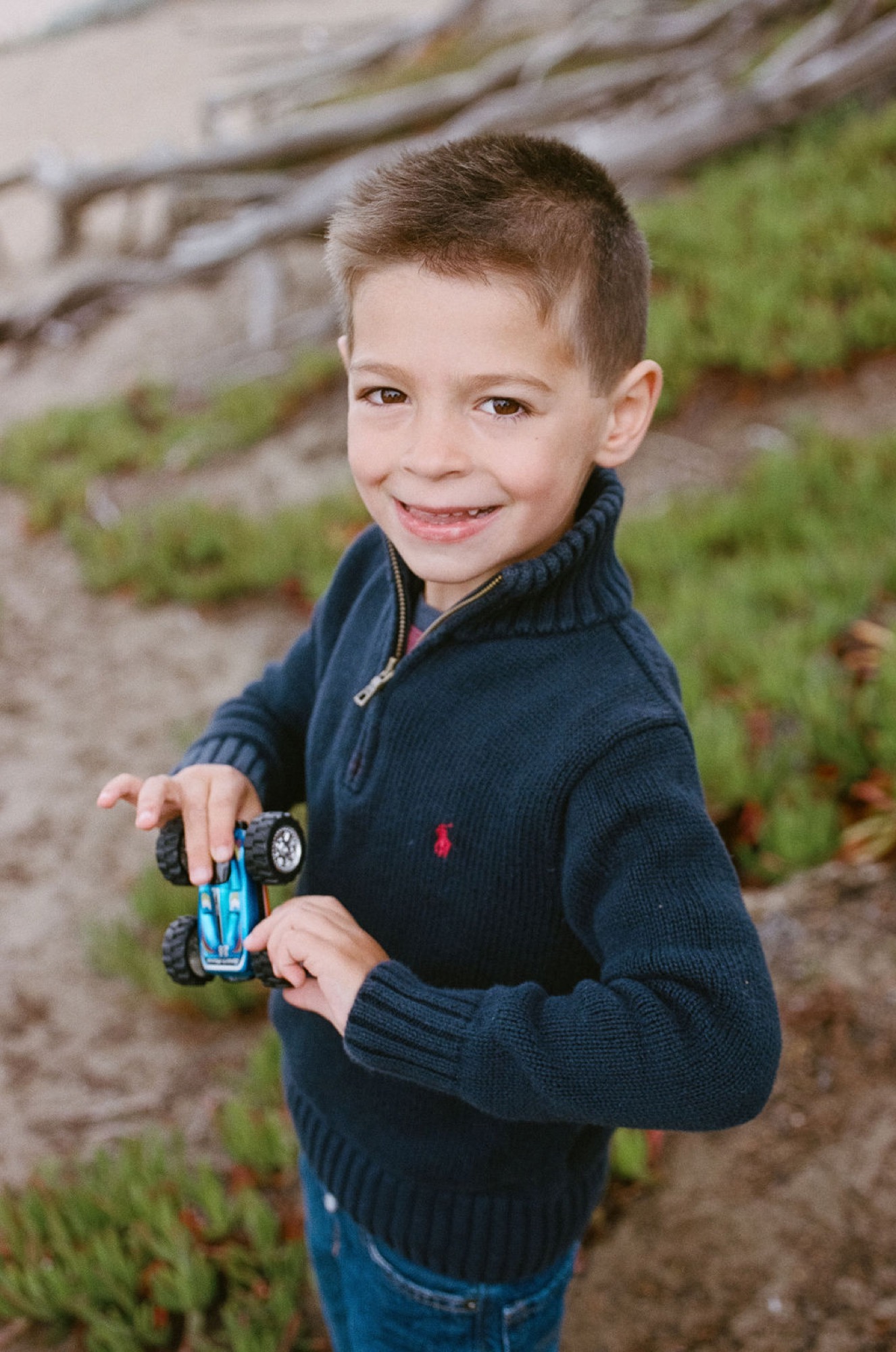 Young boy in navy zip-up smiles at camera while holding a toy car on a rocky coastal path