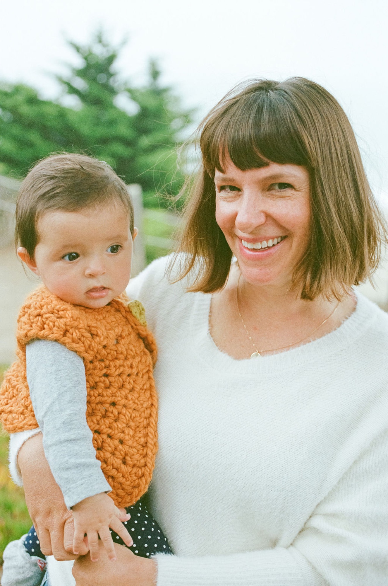 Woman in cream sweater holds smiling boy in navy cardigan with evergreen trees softly blurred behind them