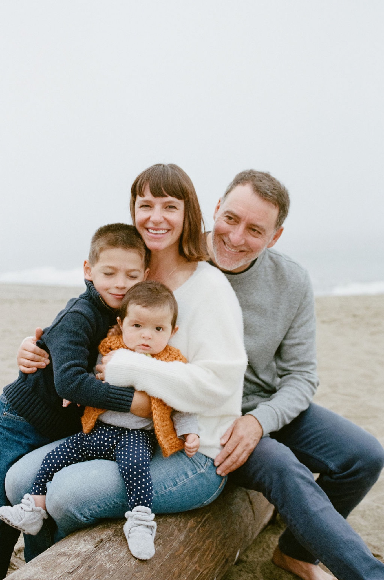 Family of four smile warmly while sitting together on driftwood, with calm beach and overcast sky behind them