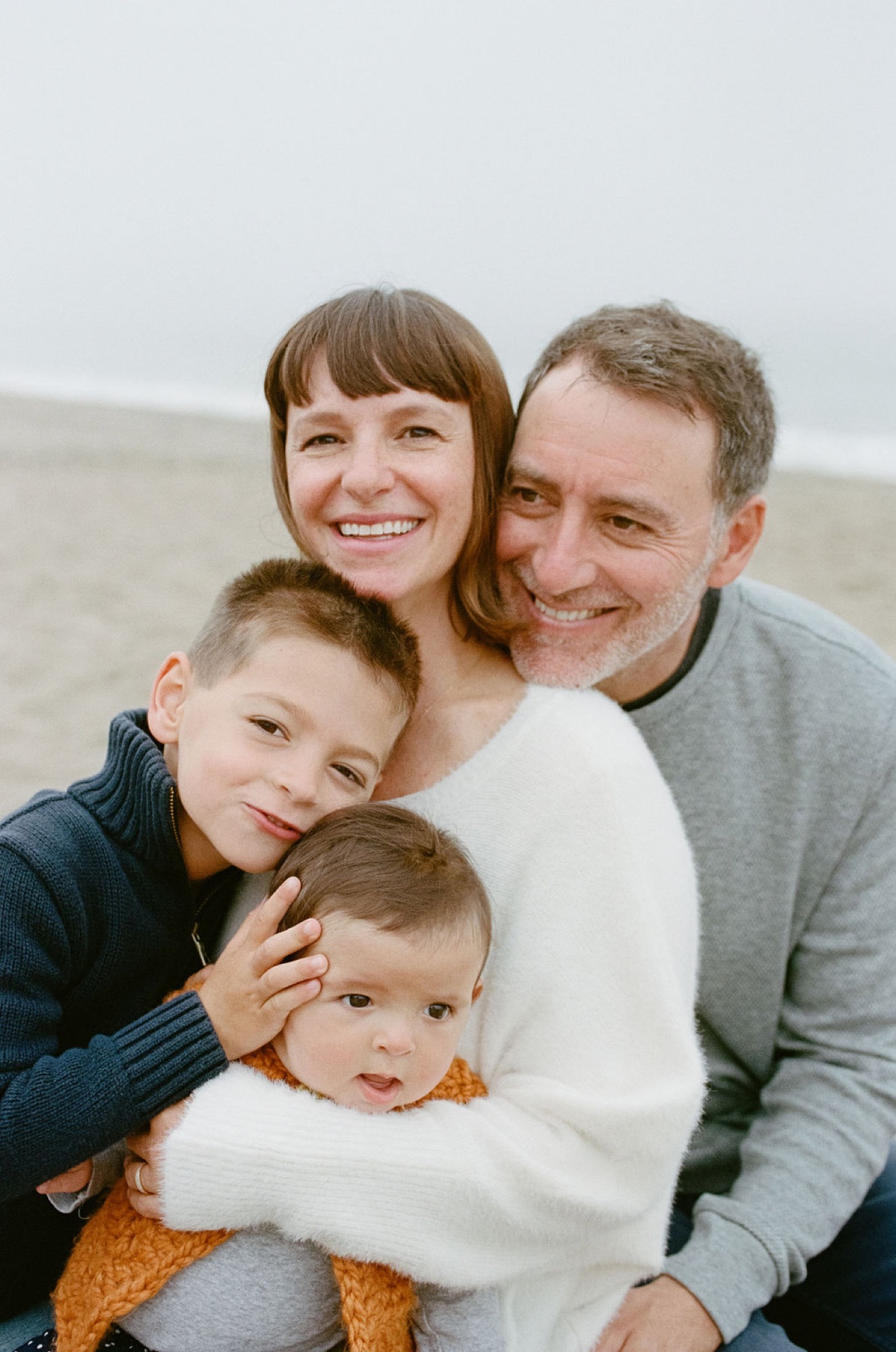 A father and mother smile warmly while embracing their two young sons on a misty beach, their genuine affection evident in their relaxed poses