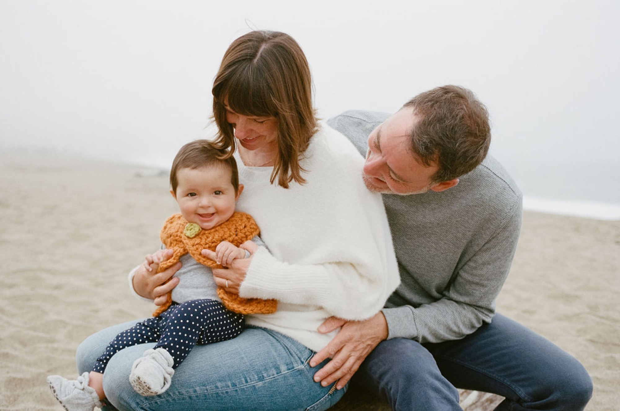 A young family kneels together on misty beach sand, the older child smiling while parents gaze down with tender affection