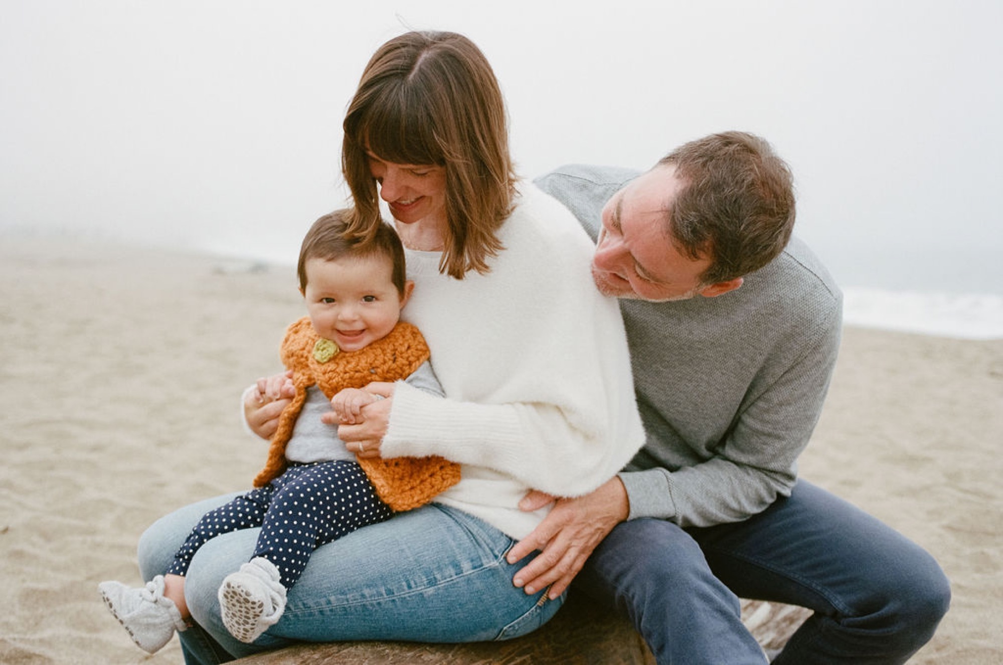 Parents kneel beside their son on misty beach sand, sharing an intimate moment of connection and joy