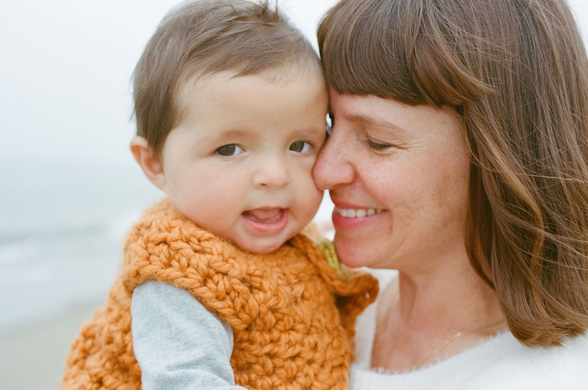A beaming mother nuzzles her toddler wearing a chunky orange sweater against a soft overcast sky