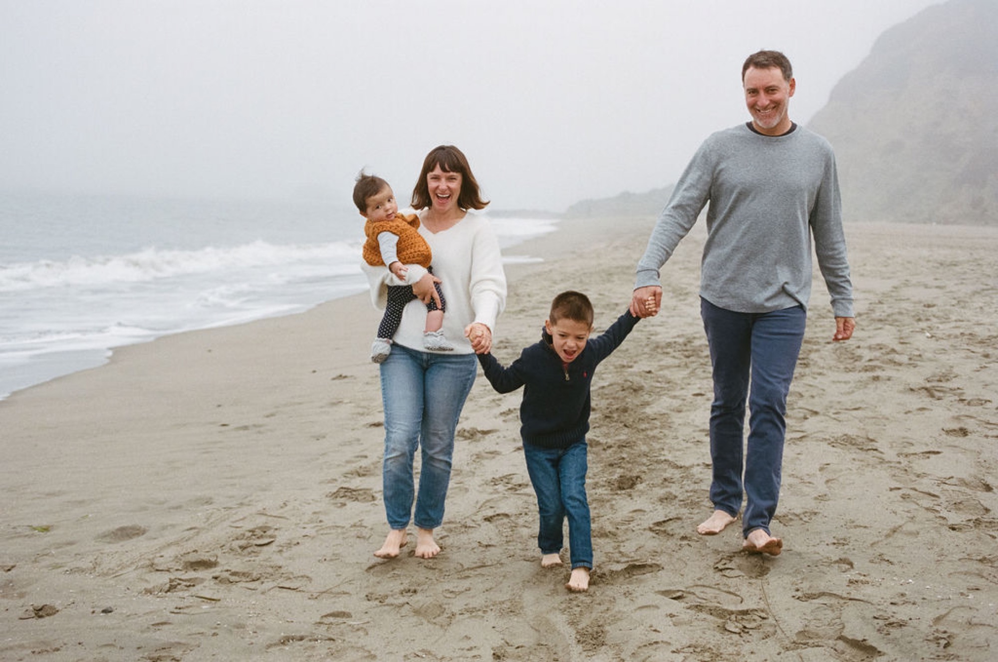Family of four walks barefoot along misty beach sand, holding hands while ocean waves crash behind them in soft overcast light