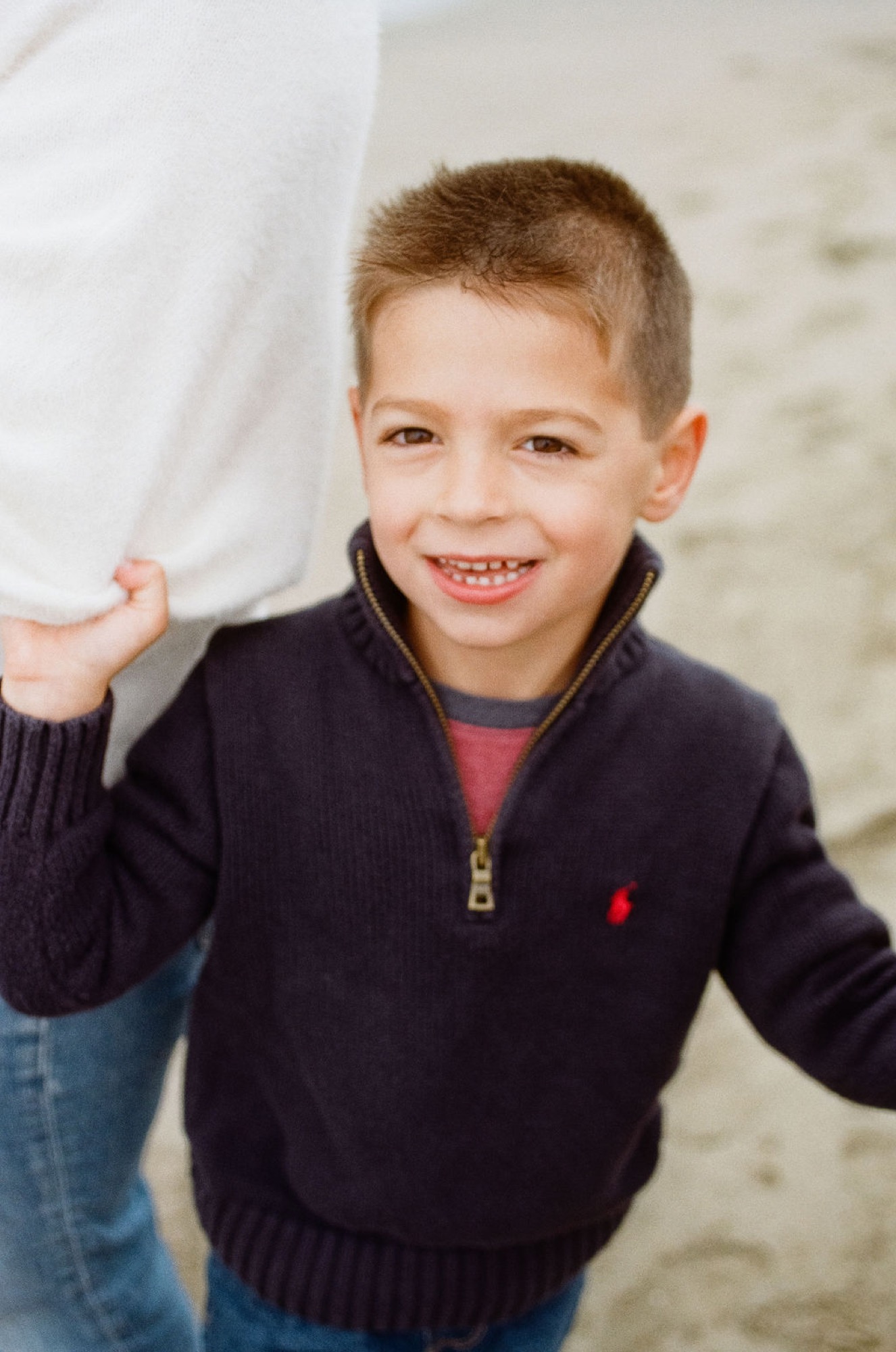 Young boy in navy sweater grins widely while holding an adult's hand at the beach, genuine joy radiating from his expression