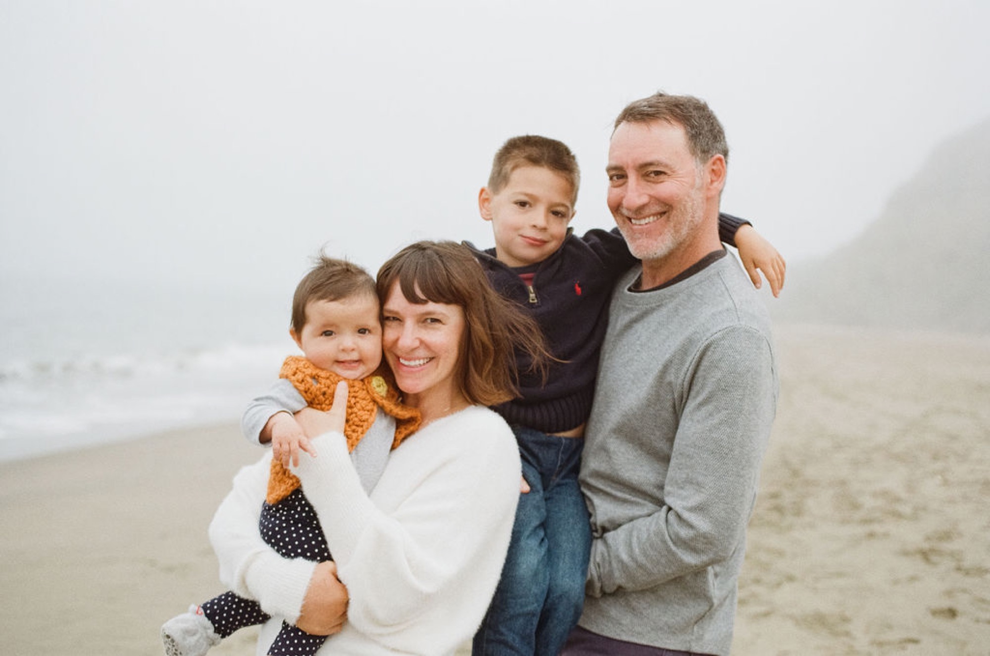 A family of four stands close together on a misty beach, with the father's arm around his older son while the mother holds a toddler, all smiling warmly at the camera