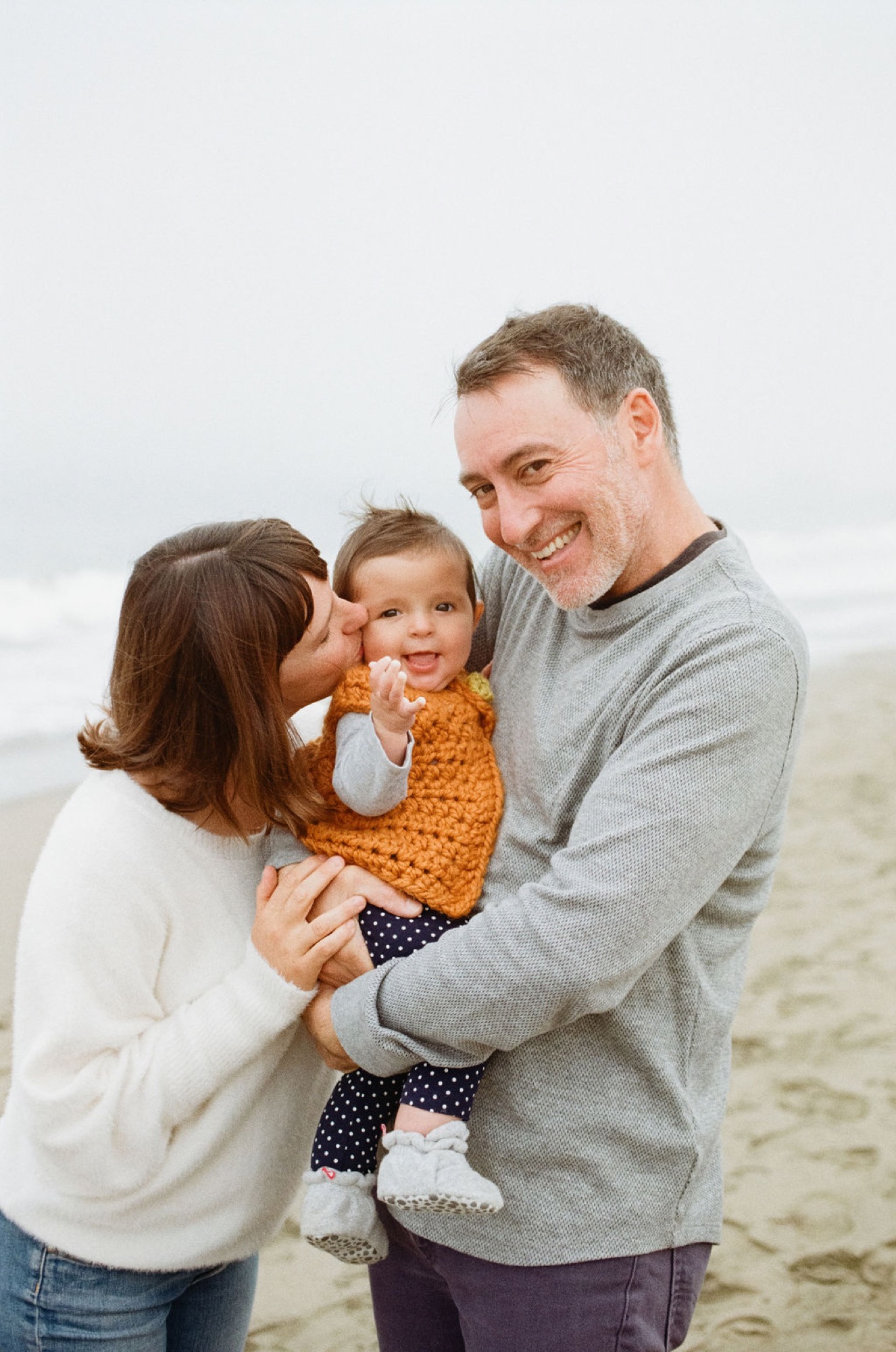 Parents kiss their toddler on the beach while holding her between them, all smiling warmly in soft overcast light
