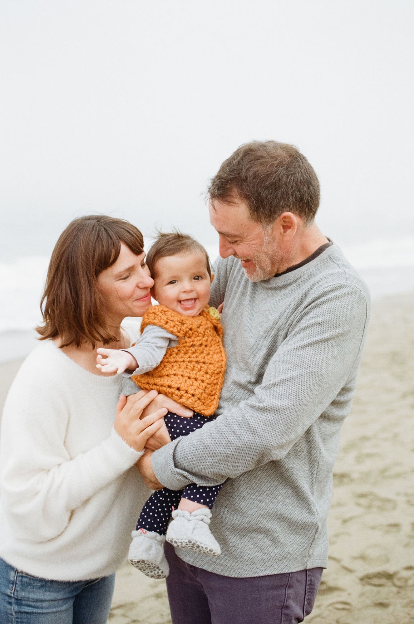Parents hold their son on an overcast beach, exchanging warm smiles while soft natural light bathes the sandy shore