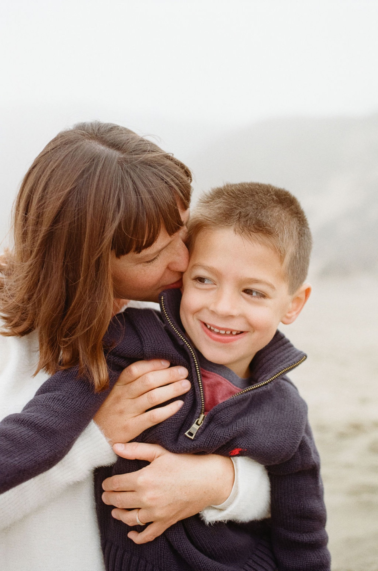 A young boy grins at the camera while his older sister kisses his cheek, both wearing dark hoodies against a soft, overcast beach backdrop