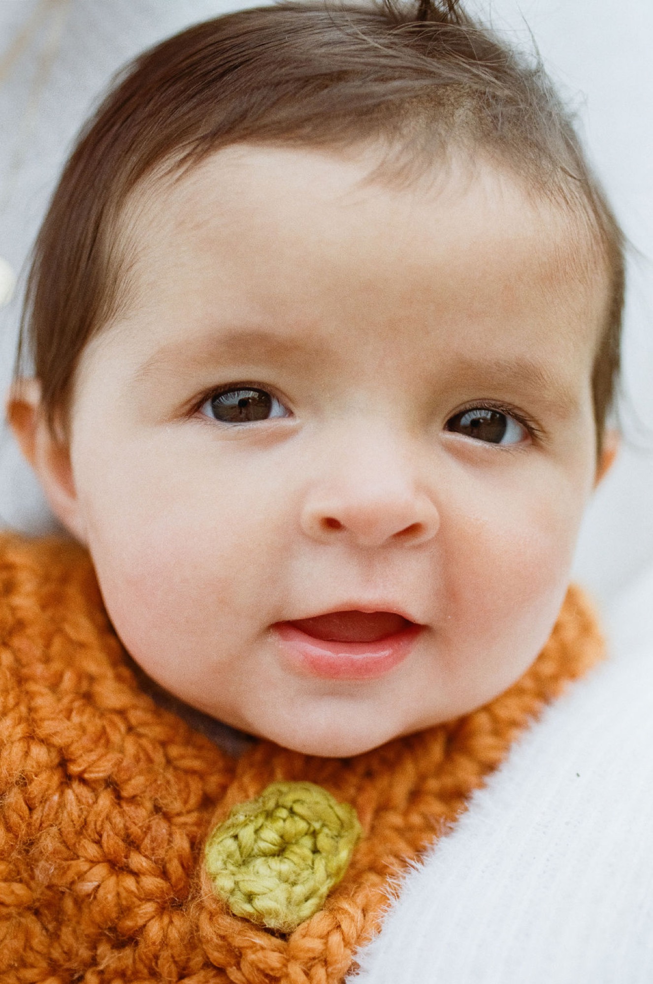 Baby girl in golden-yellow knit sweater gazes directly at camera with wide, alert eyes and slight smile