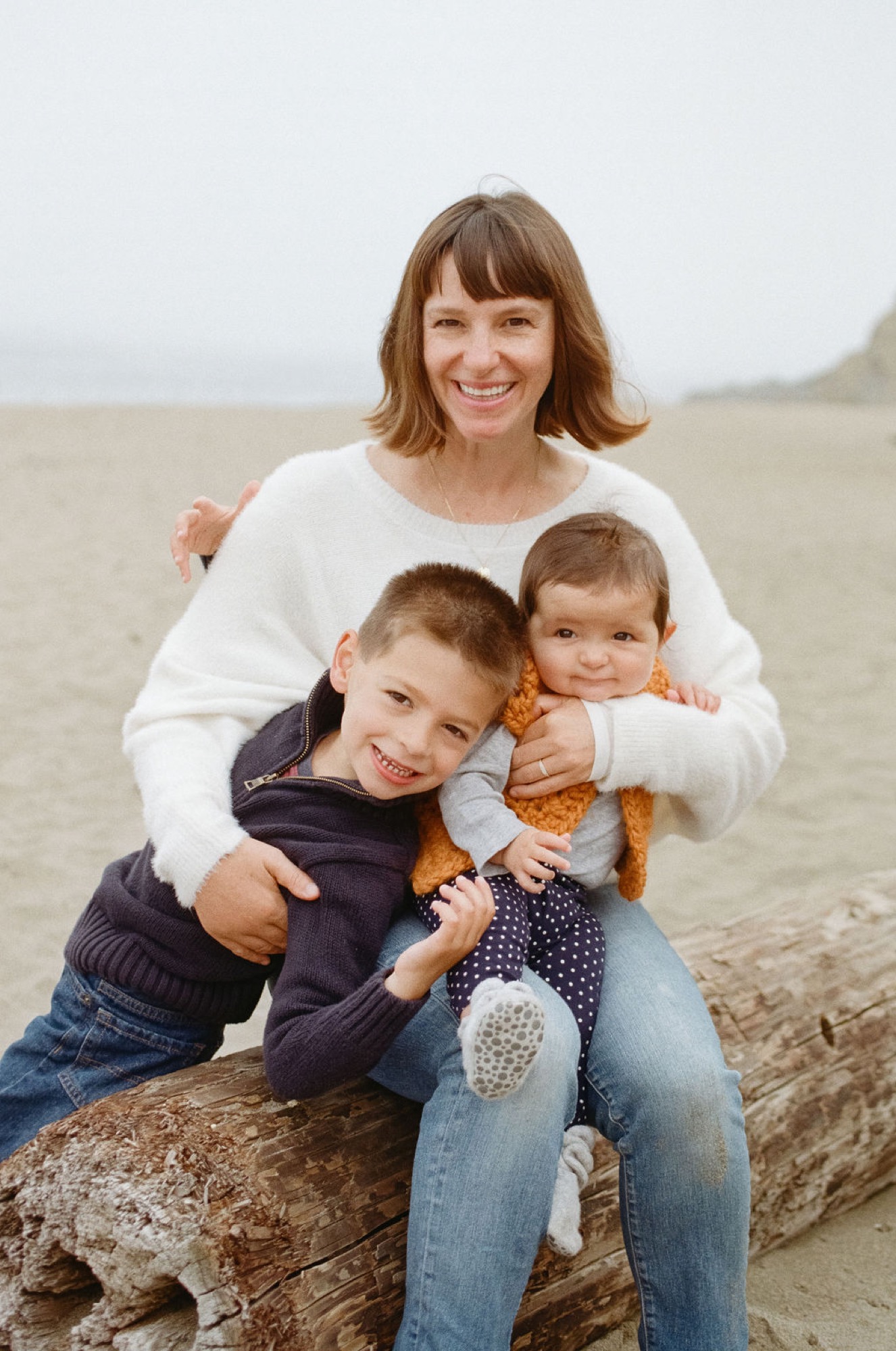 Woman and two young boys smile warmly while sitting on driftwood, ocean and beach stretching behind them in soft natural light