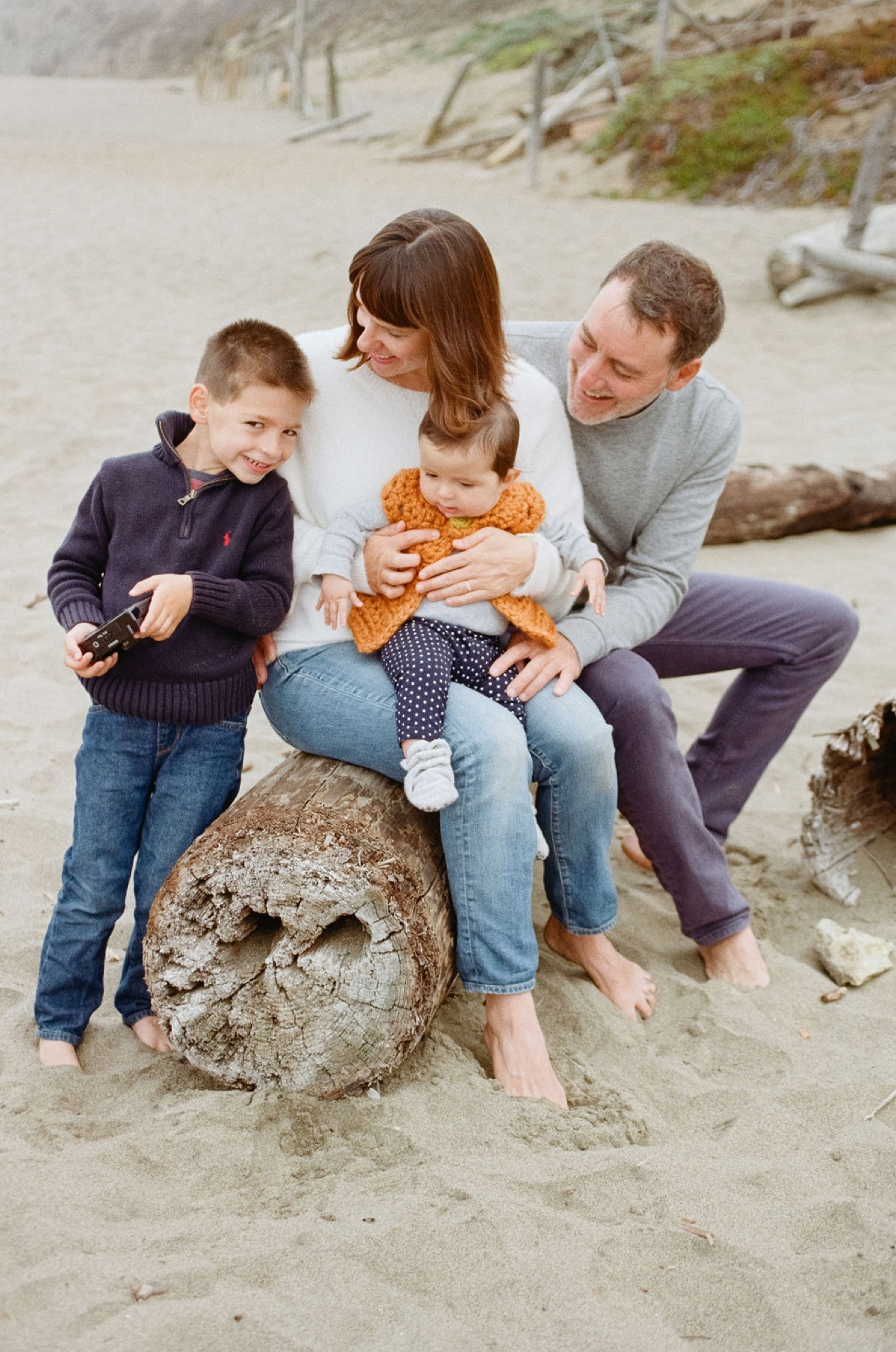 Two adults and two young children sit together on a weathered log at the beach, sharing a relaxed moment in natural light