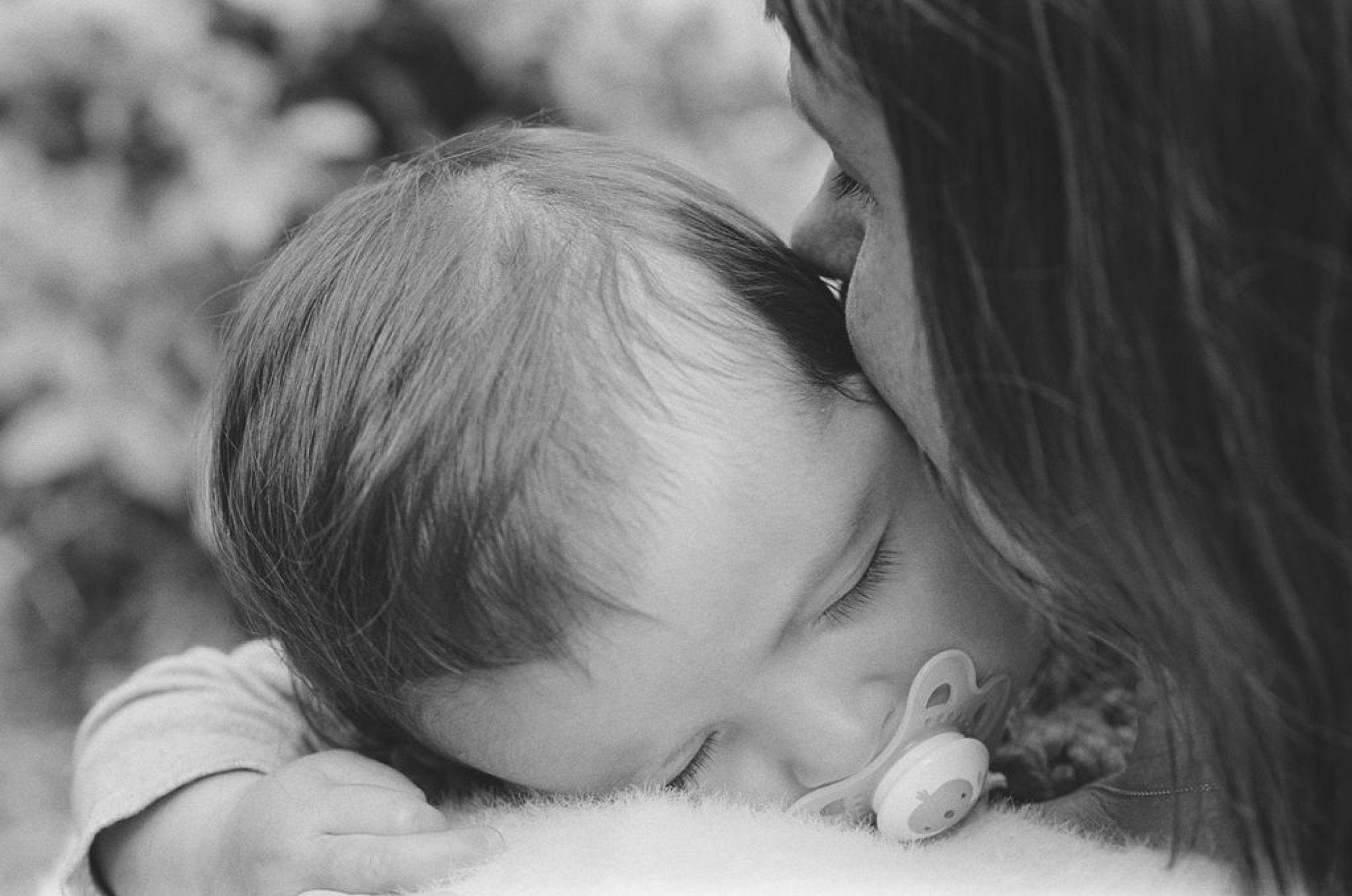A sleeping baby girl nestles against a parent's shoulder in a tender black and white moment, soft natural light catching delicate wisps of baby hair