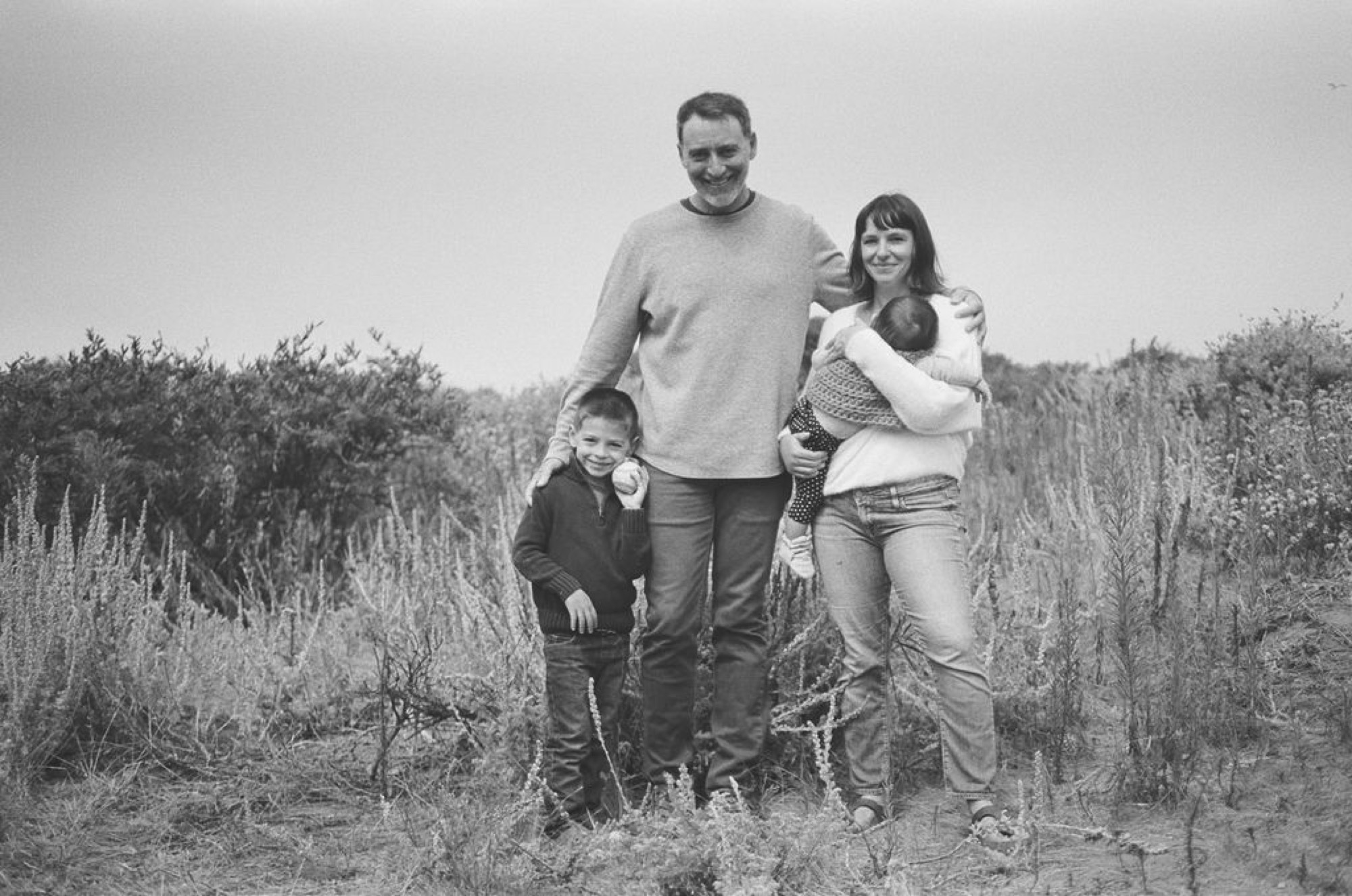 A family of four stands together on a hillside path, the adults flanking two children with gentle expressions in soft, diffused light