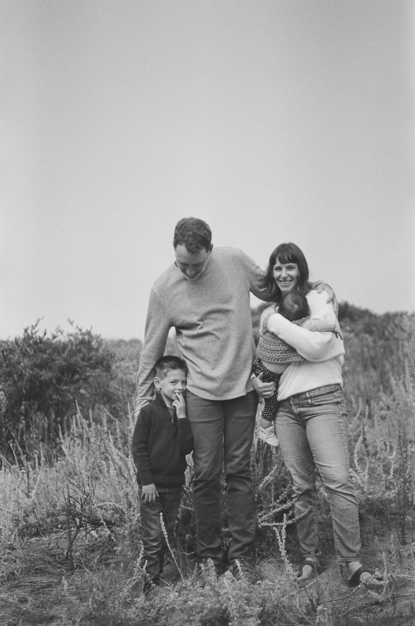 Three figures walk together through tall grass on a hillside, the adult looking down warmly while the child gazes ahead in this intimate black and white moment