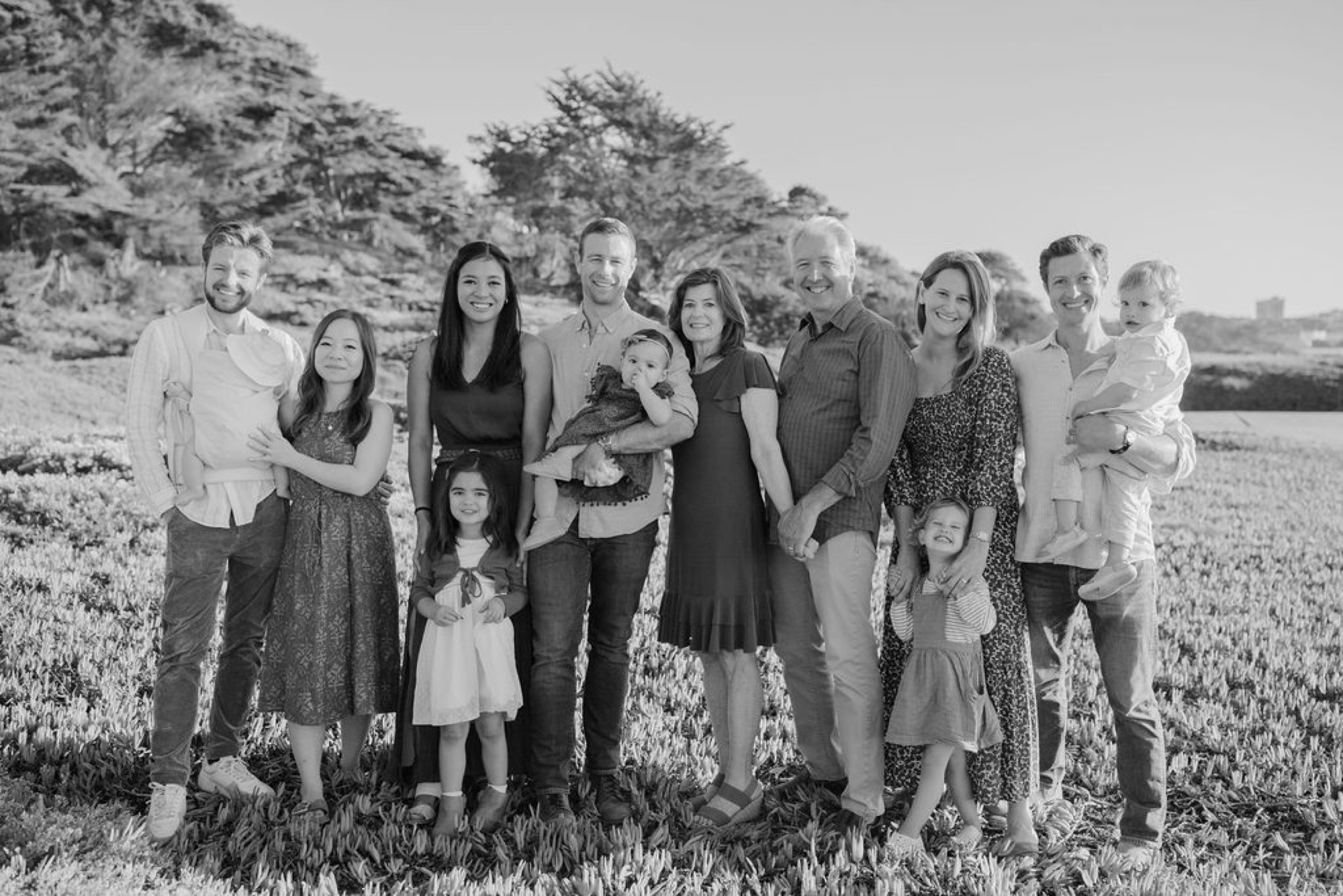 A multi-generational family stands shoulder-to-shoulder in an open meadow, their relaxed postures and gentle expressions captured in warm, diffused natural light