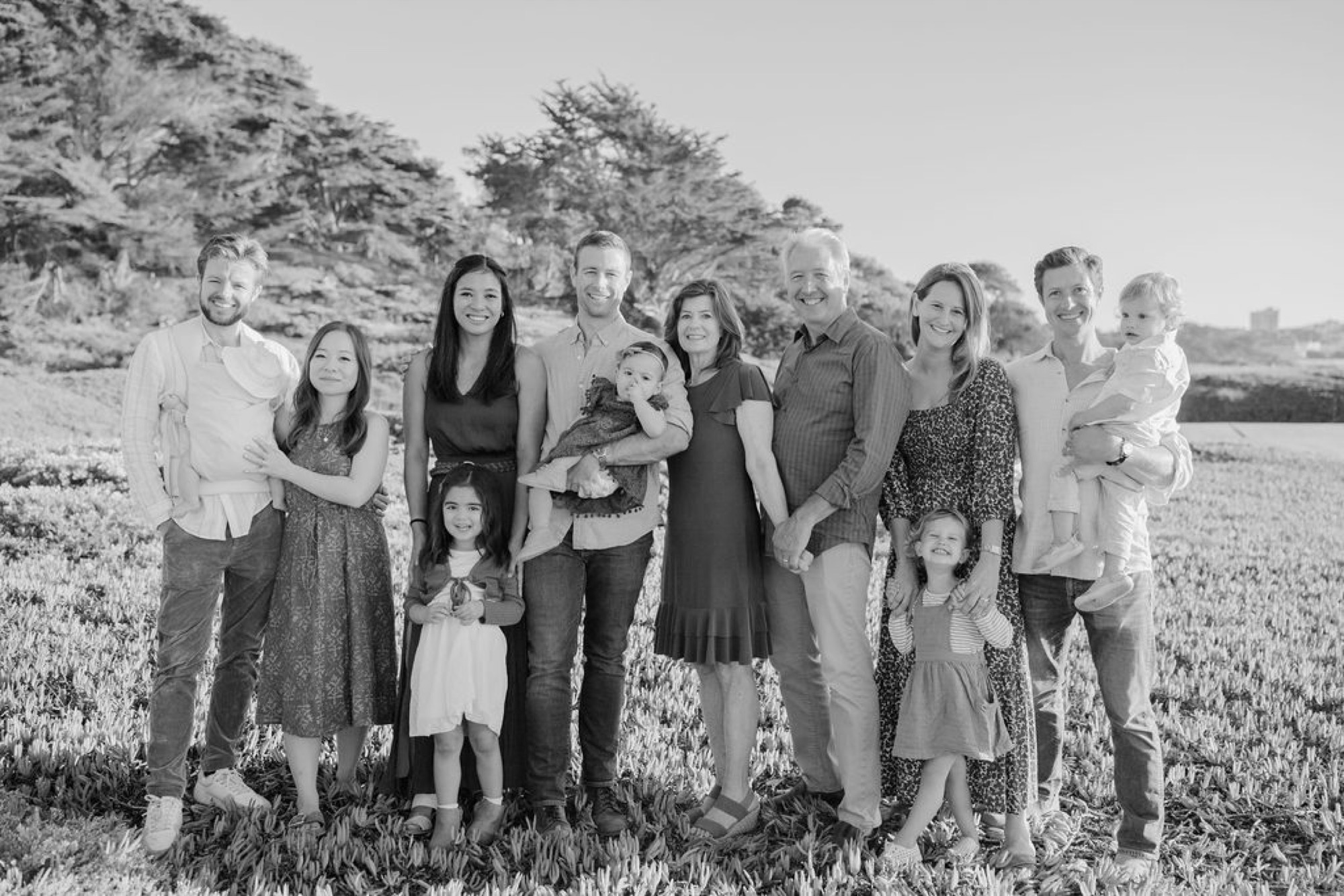 Large multigenerational family stands closely together on rolling grassland, many smiling toward camera, with windswept trees and open fields behind them