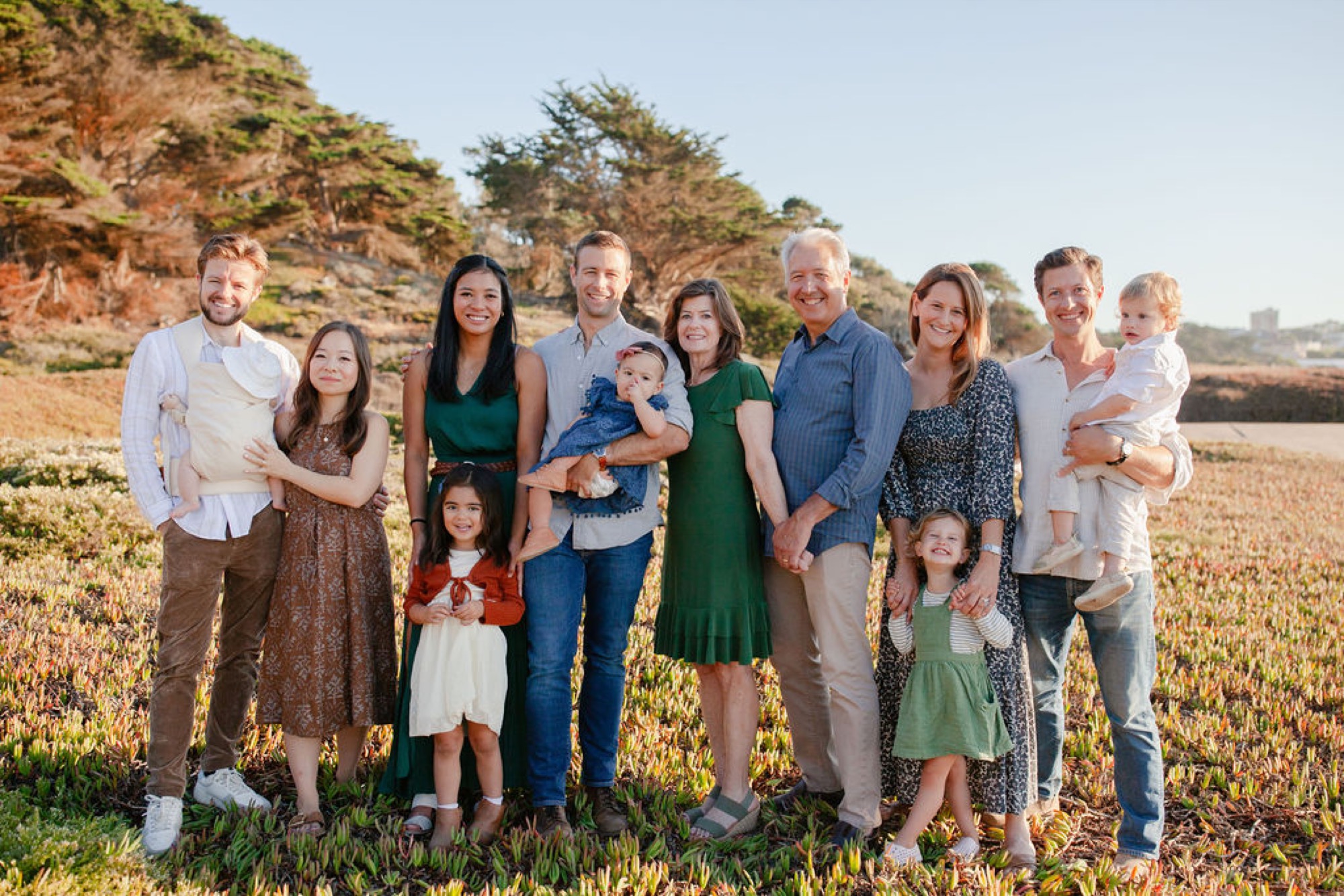 Three-generation family stands together on a sunlit hillside with golden grass and coastal scrub, wearing earth tones and pastels in relaxed poses