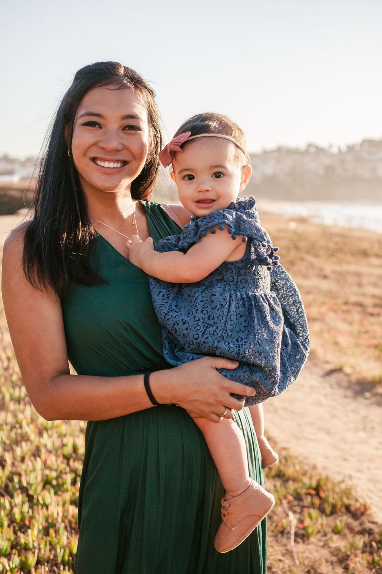 Woman in emerald dress holds smiling toddler wearing headband on sun-drenched beach with blurred coastal hills behind