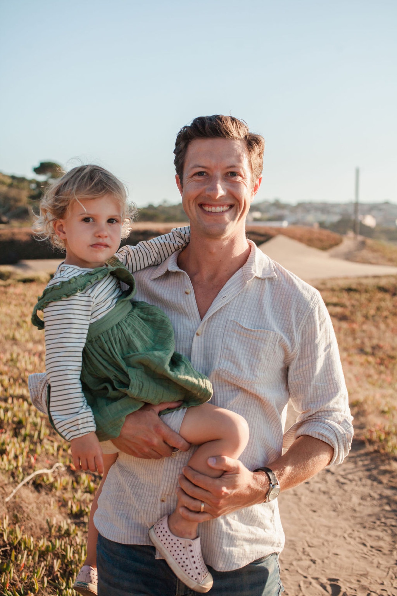 A smiling father holds his young son in a striped shirt across a sunlit dirt road with rolling hills and sparse vegetation behind them
