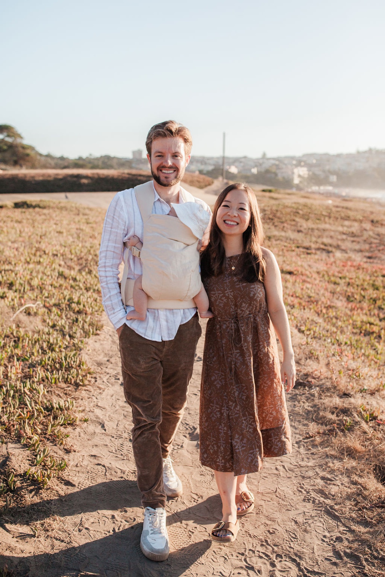 A smiling couple walks hand-in-hand through a harvested golden field under warm afternoon sunlight, hills visible in the distant background