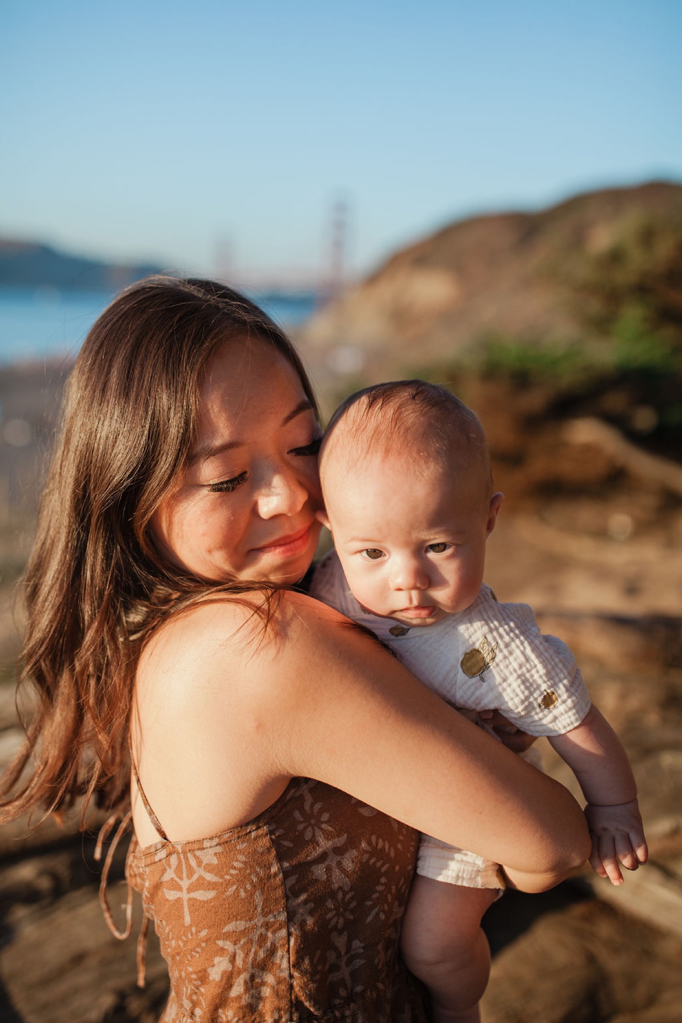 A woman holds her toddler on rocky terrain with the Golden Gate Bridge visible in the distant hazy background