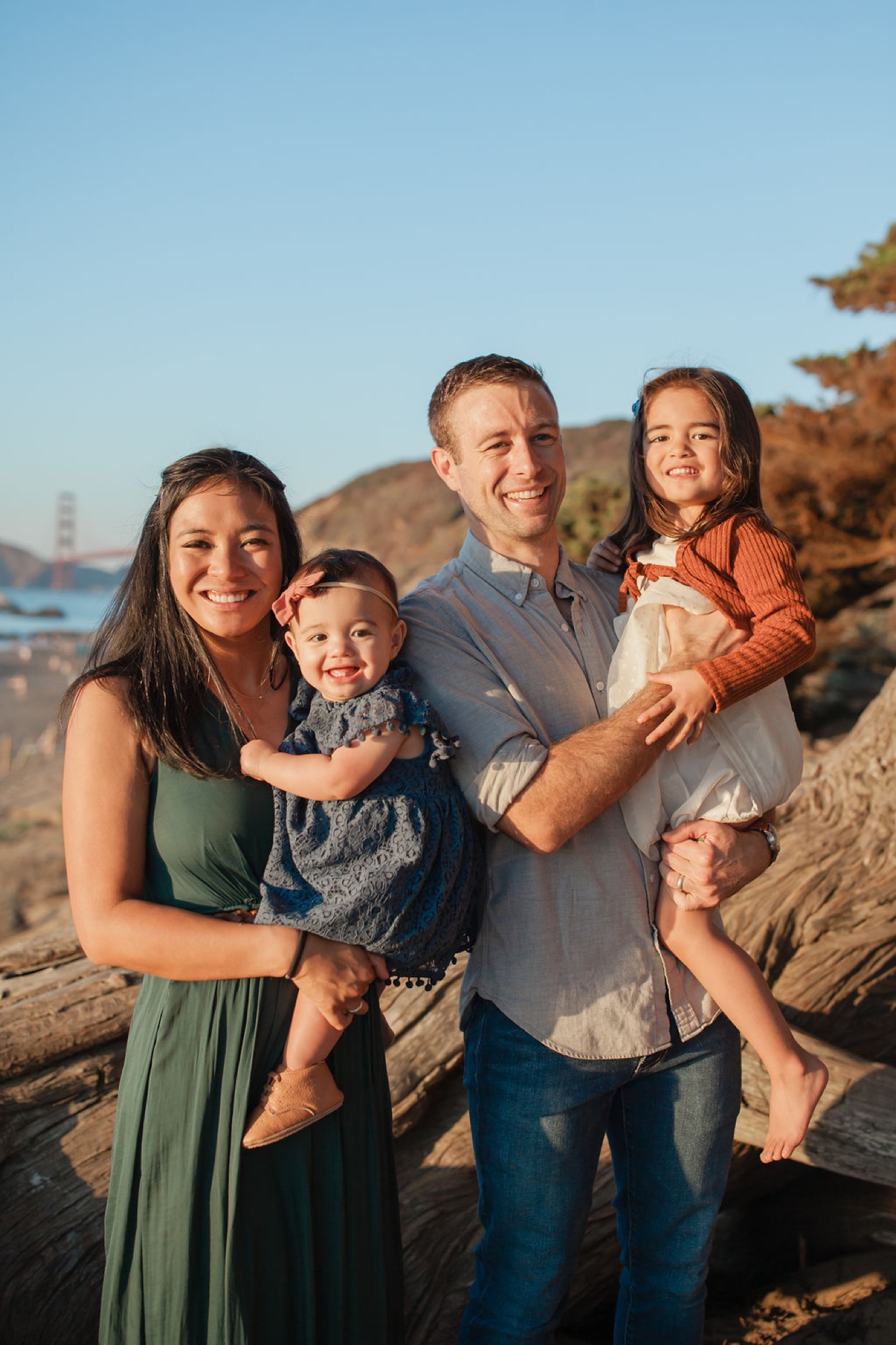Family of five smiles warmly at sunset on rocky Baker Beach, with golden light illuminating their relaxed poses and genuine expressions