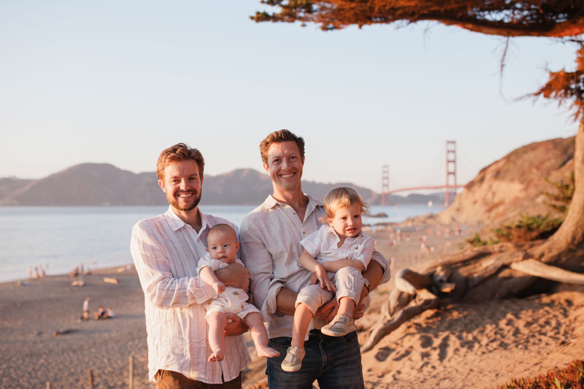 Two fathers smile warmly while holding their young children on a sandy beach, the Golden Gate Bridge glowing in the soft afternoon light behind them