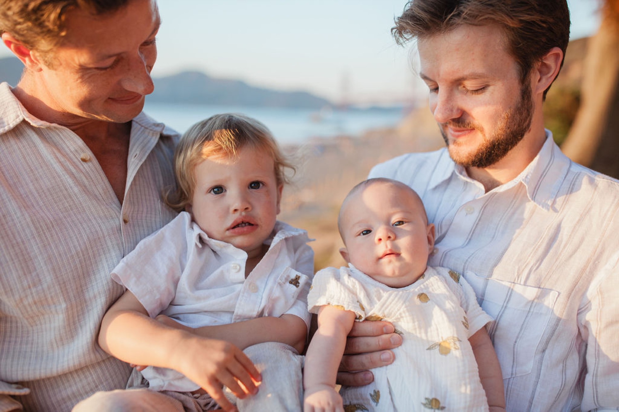 Two men hold toddlers at sunset on a beach, their expressions tender and focused downward toward the children in soft golden light