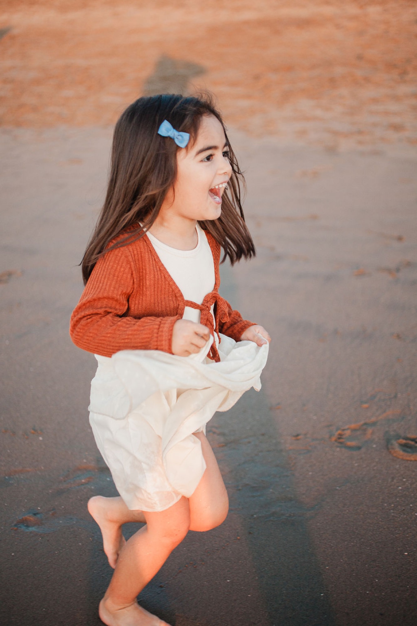 Young girl running barefoot on wet sand with wind-tousled hair, wearing rust cardigan and cream skirt against golden hour light