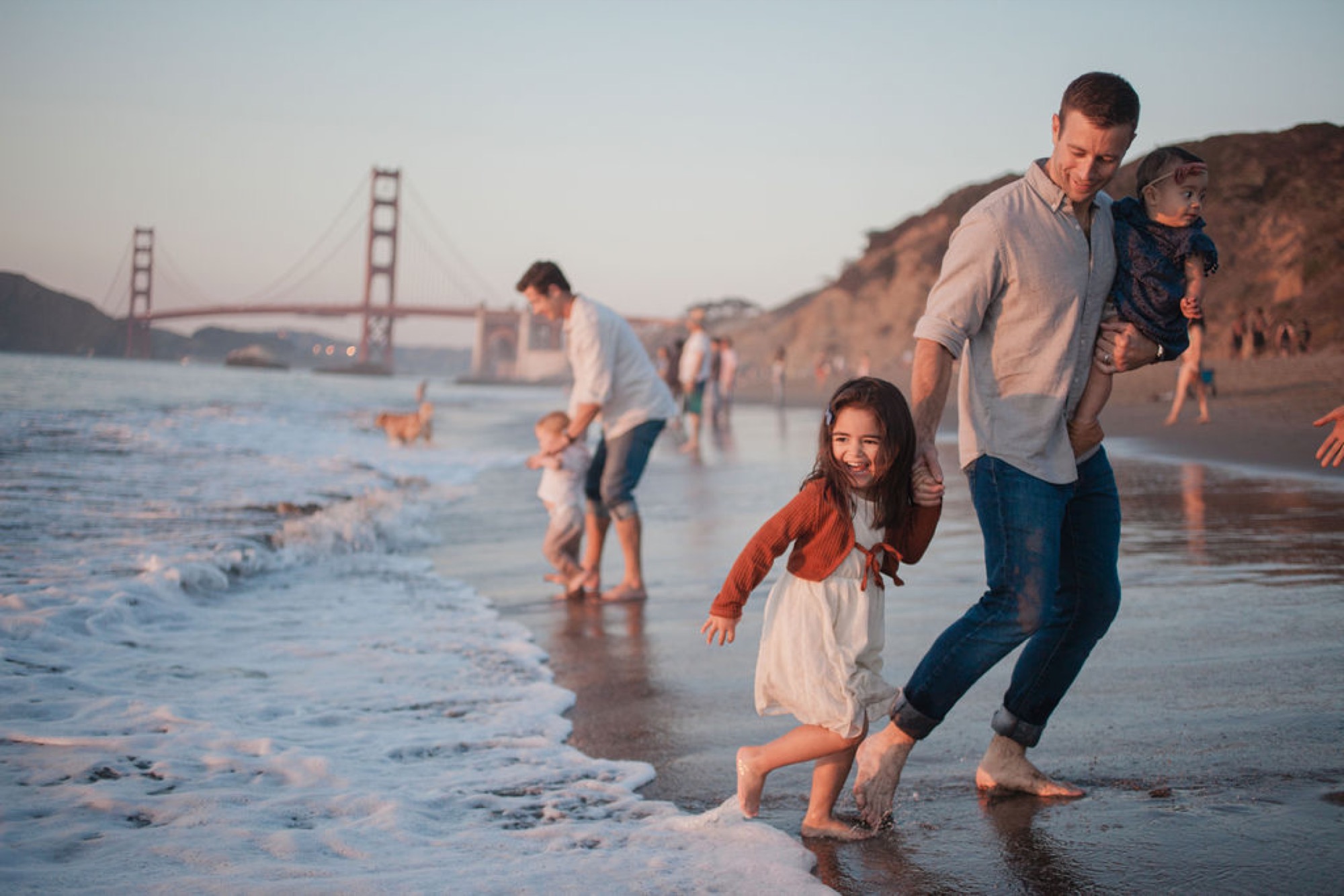 A father carries his young daughter while walking through shallow waves at Baker Beach, with the Golden Gate Bridge glowing in the golden hour light behind them