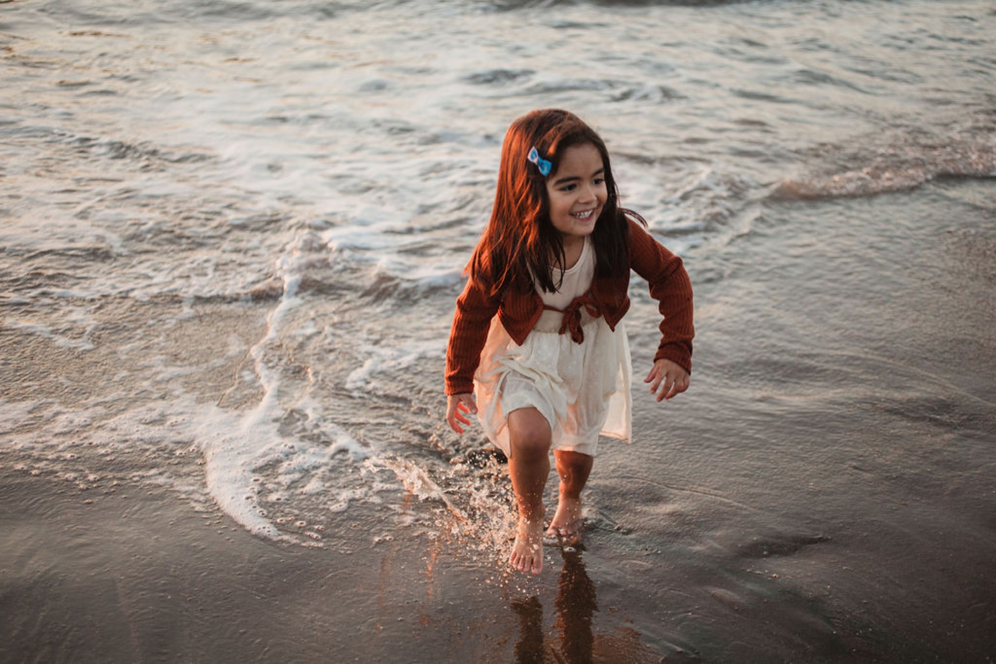 Girl with flowing red hair wades through shallow waves at sunset, glancing back with a gentle expression as golden light catches the water