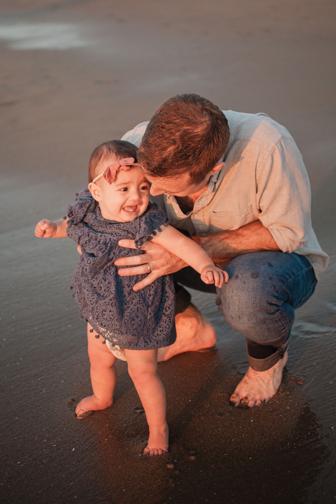 A man crouches to kiss a toddler's cheek on wet sand as golden hour light bathes their figures