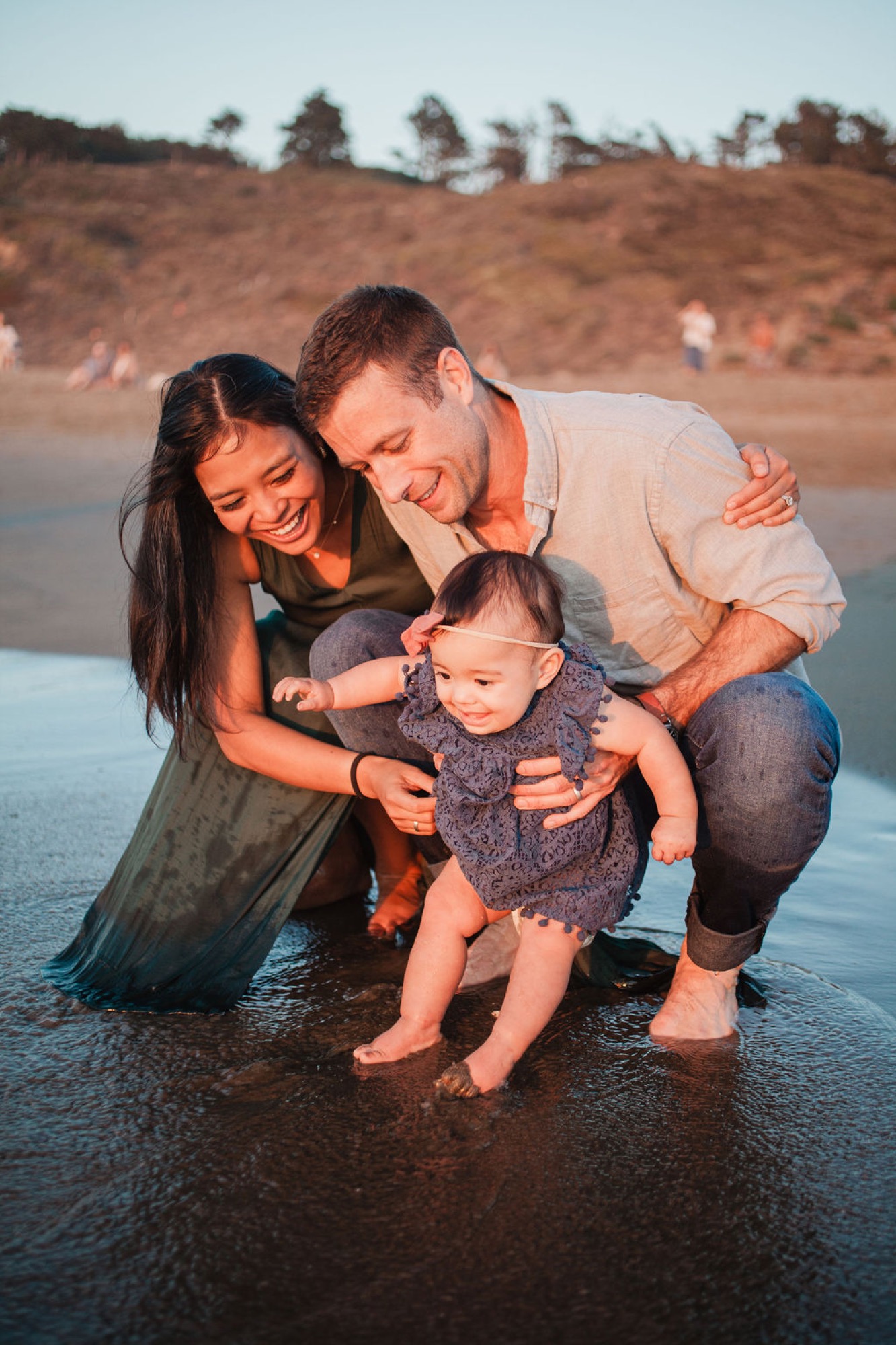 A young family crouches together in shallow tide pools at sunset, their warm silhouettes framed against golden hillsides