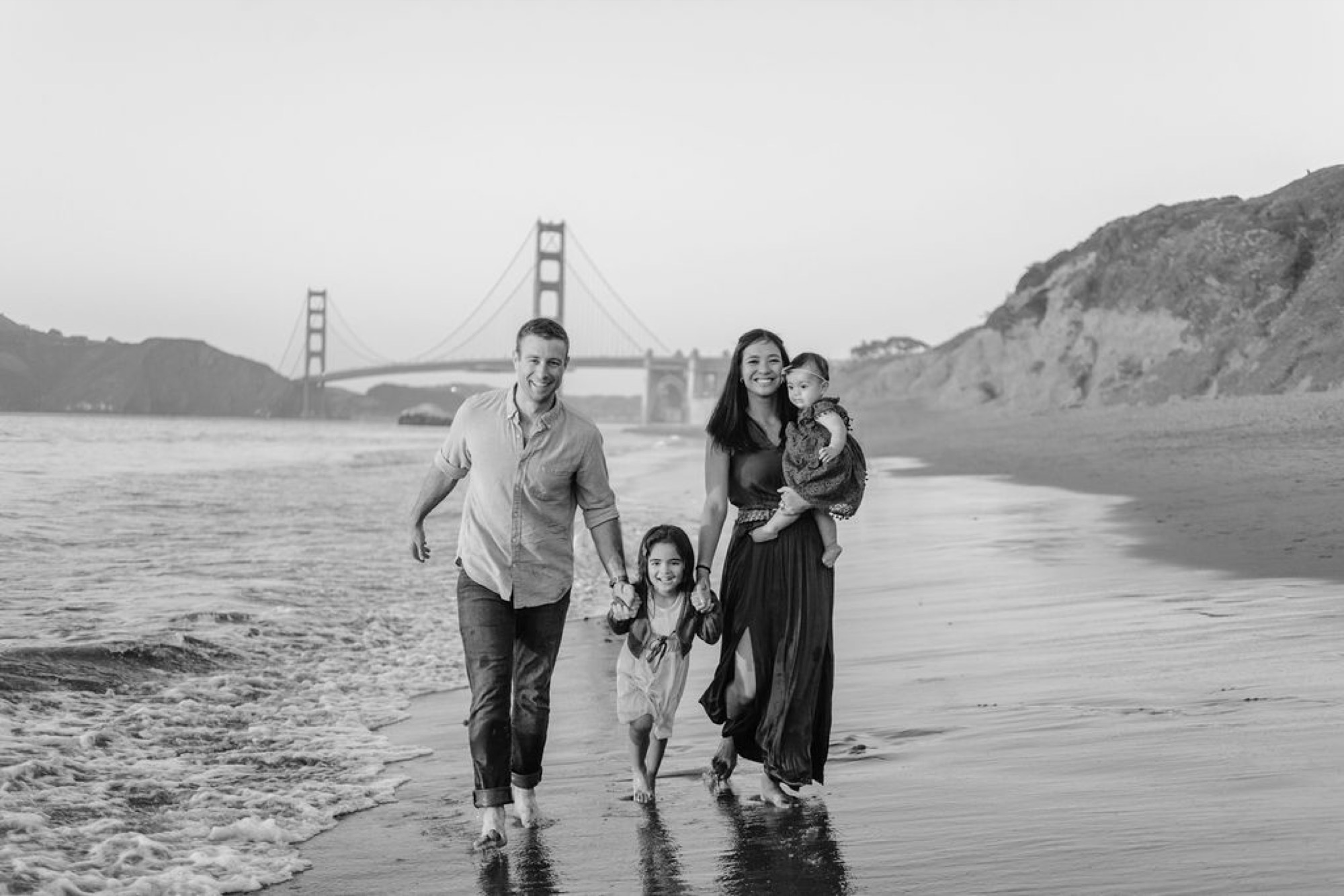 A family of five walks together through shallow water at Baker Beach, the Golden Gate Bridge visible in misty background