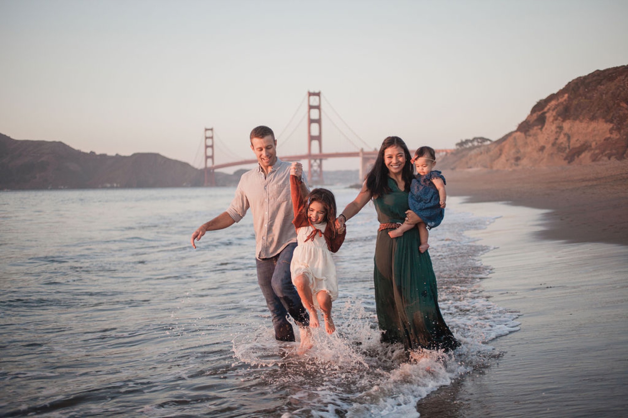 Family splashes through shallow waves at sunset with the Golden Gate Bridge glowing in the distance behind them