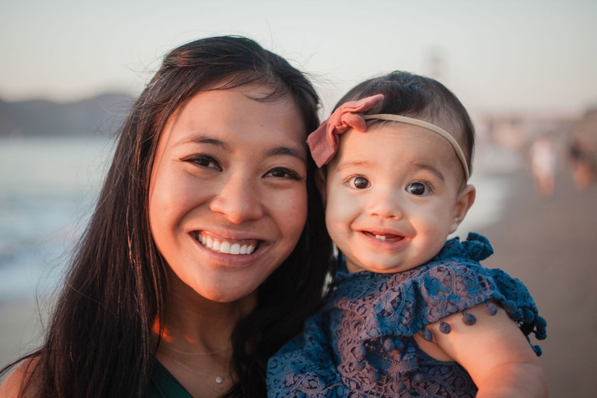 An older sister and toddler share genuine smiles at sunset, with water and distant shoreline softly blurred behind them